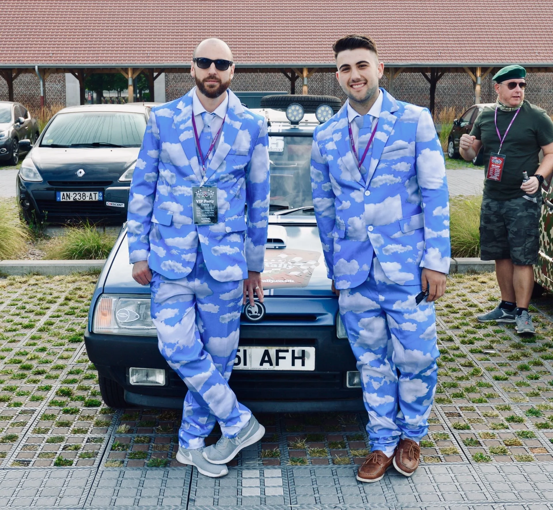 Two men in matching blue and white cloud-patterned suits stand in front of a vintage black car with a license plate reading 51 AFH, smiling at the camera. They are surrounded by parked cars and other people in the background, one of whom is wearing a