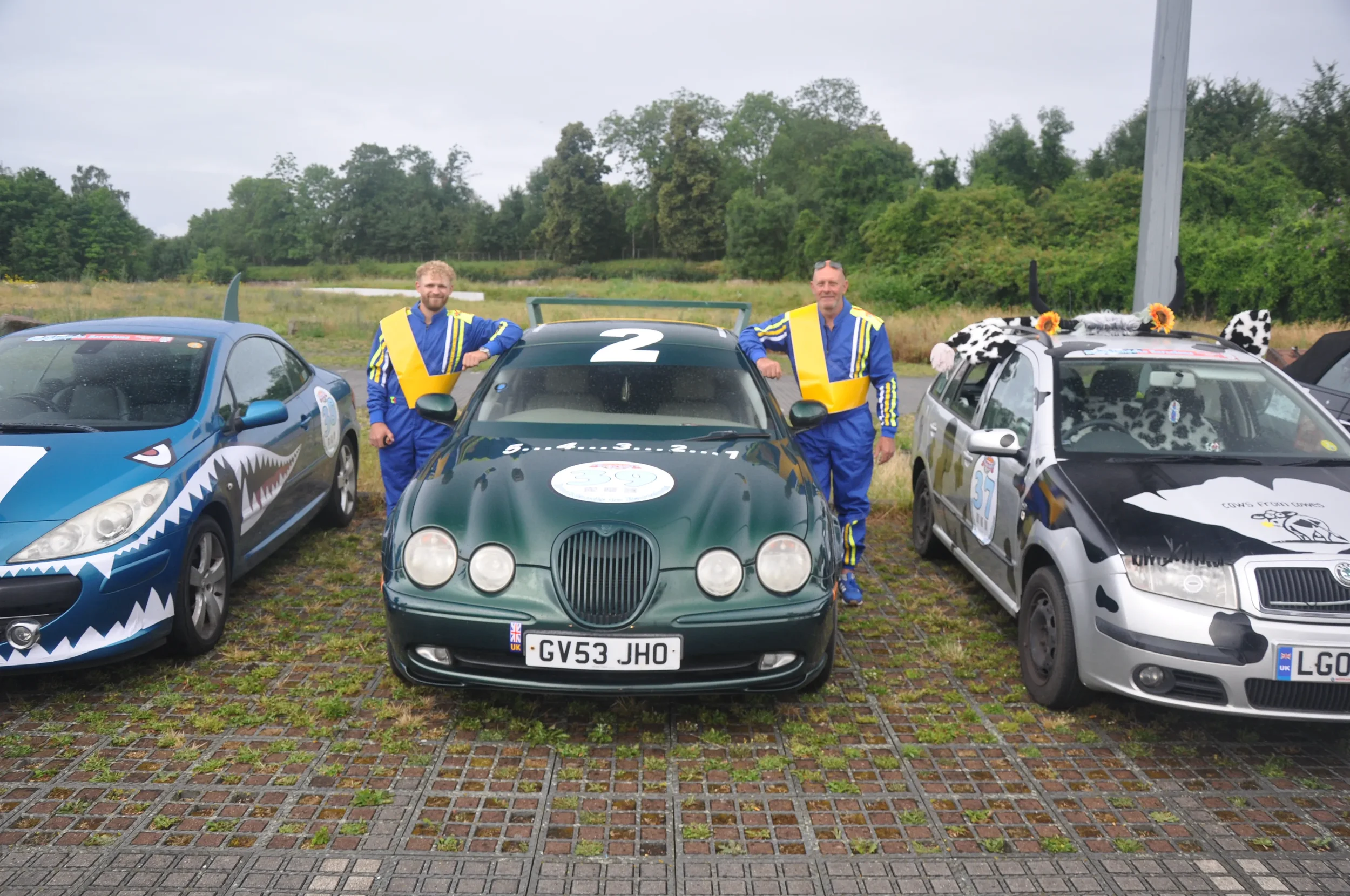 Three race cars with people in racing suits standing beside them in an outdoor parking lot with grassy field and trees in the background.