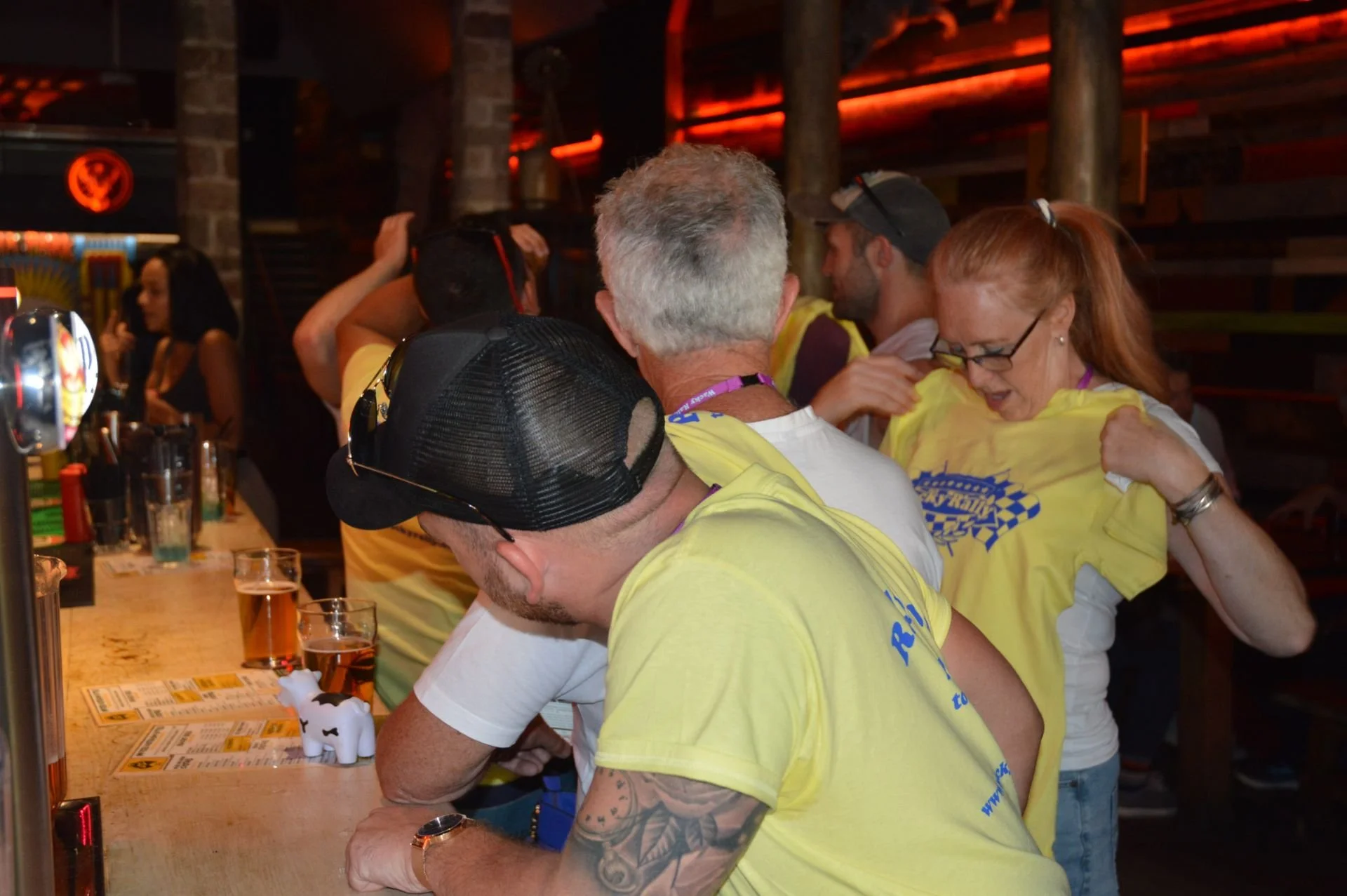 People wearing yellow shirts at a bar, engaging in conversation and activities, with drinks and a cow figurine on the counter.