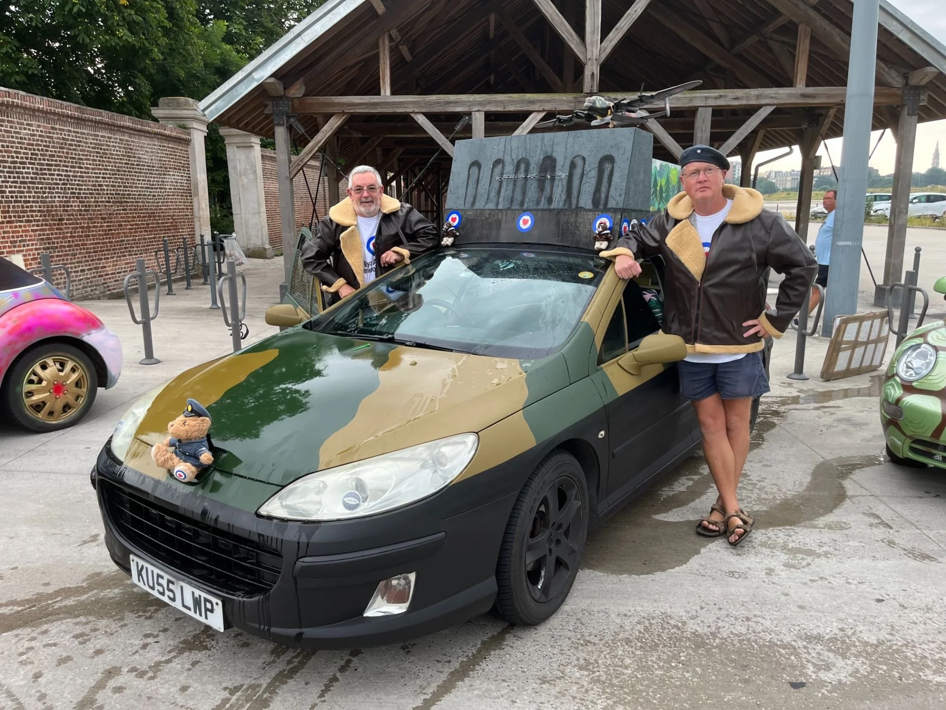 Two men wearing leather jackets with shearling collars posing beside a camouflage-painted car with a teddy bear dressed as a pilot on its hood. The car is parked in an outdoor area with other colorful toy cars and a wooden pavilion.