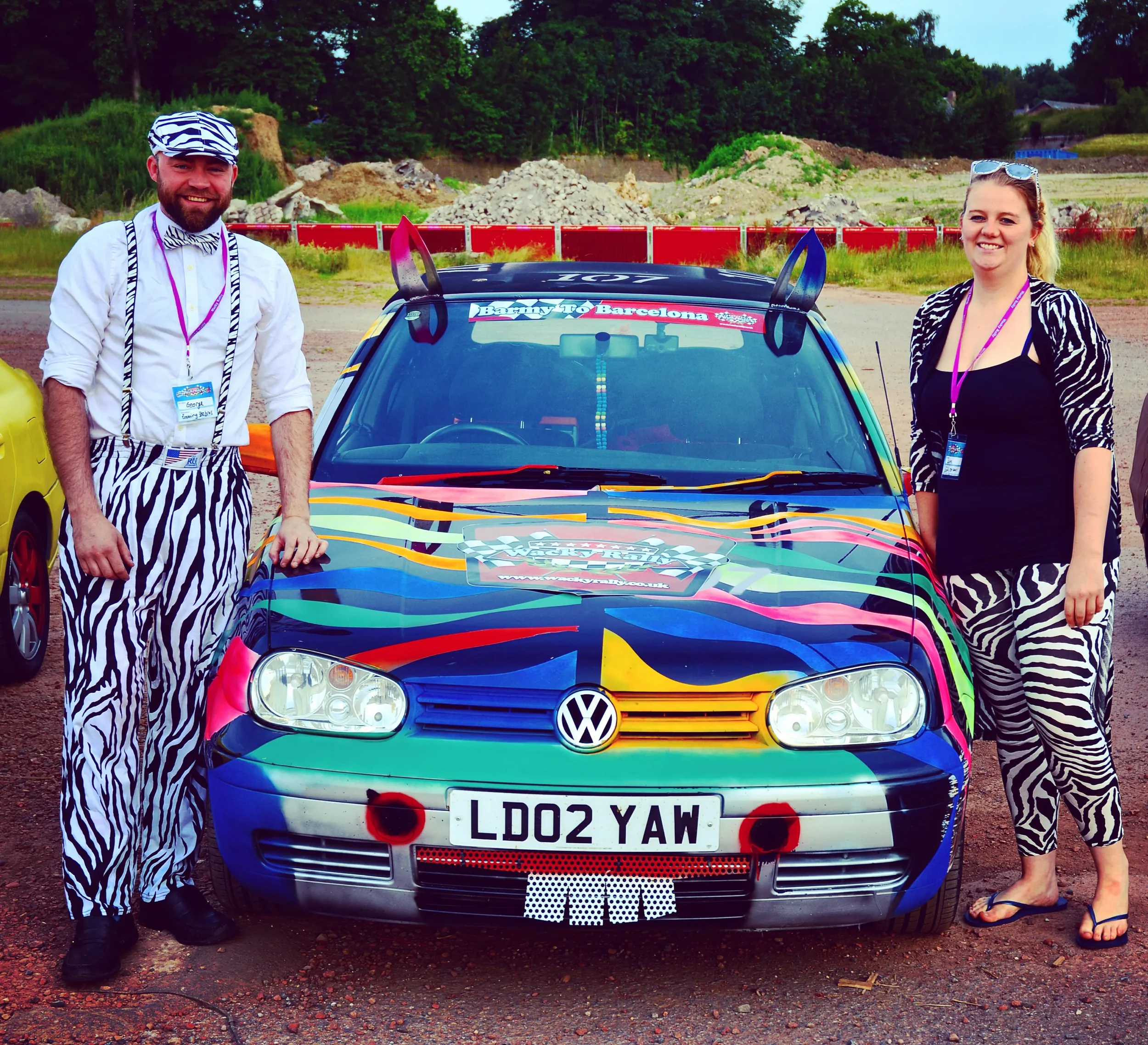 Two people dressed in zebra print outfits standing next to a colorful Volkswagen car with a custom paint job, at an outdoor event with trees and construction materials in the background.