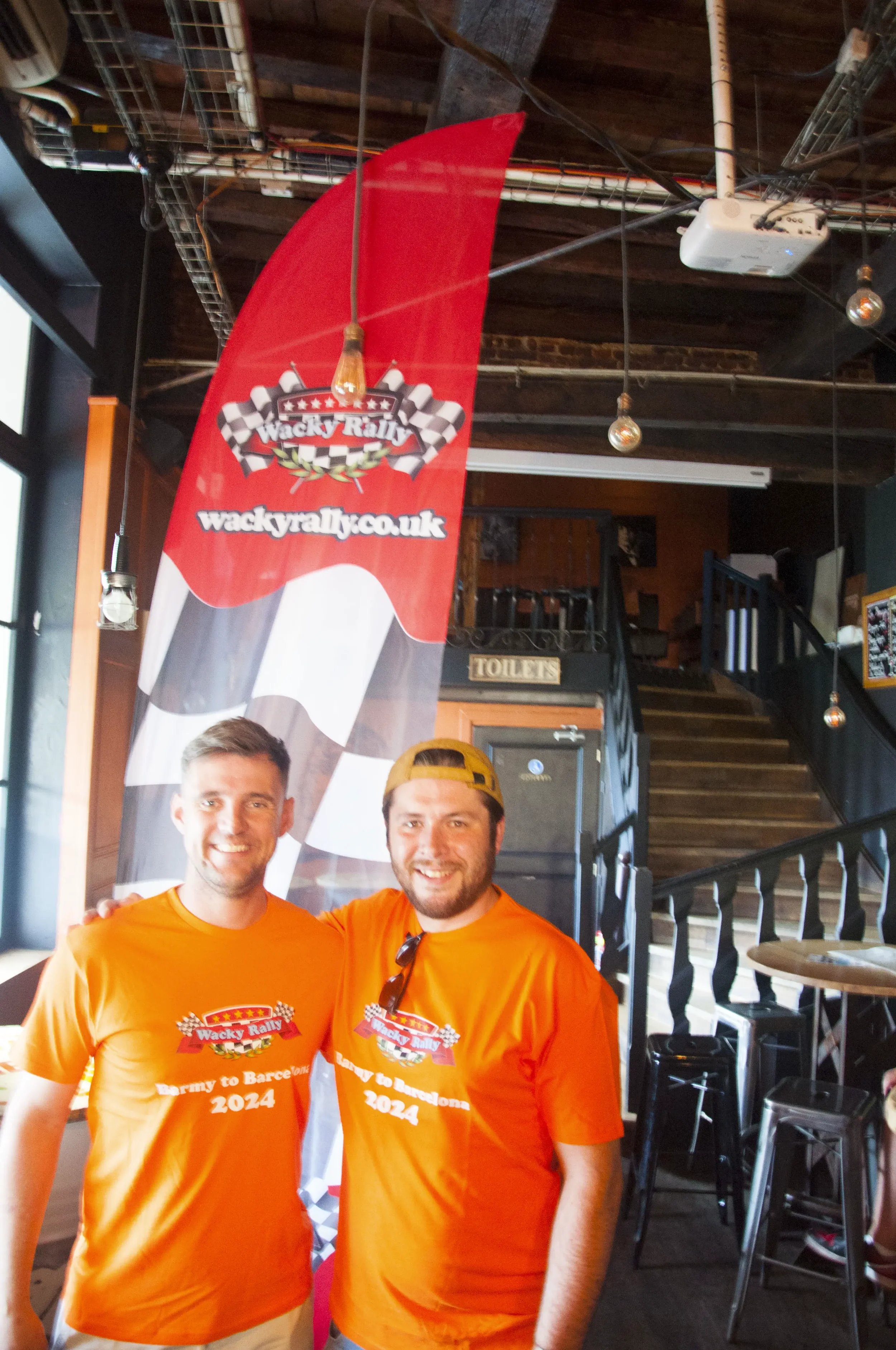 Two smiling men wearing orange Wacky Rally event shirts, standing inside a venue with a large red Wacky Rally banner behind them.