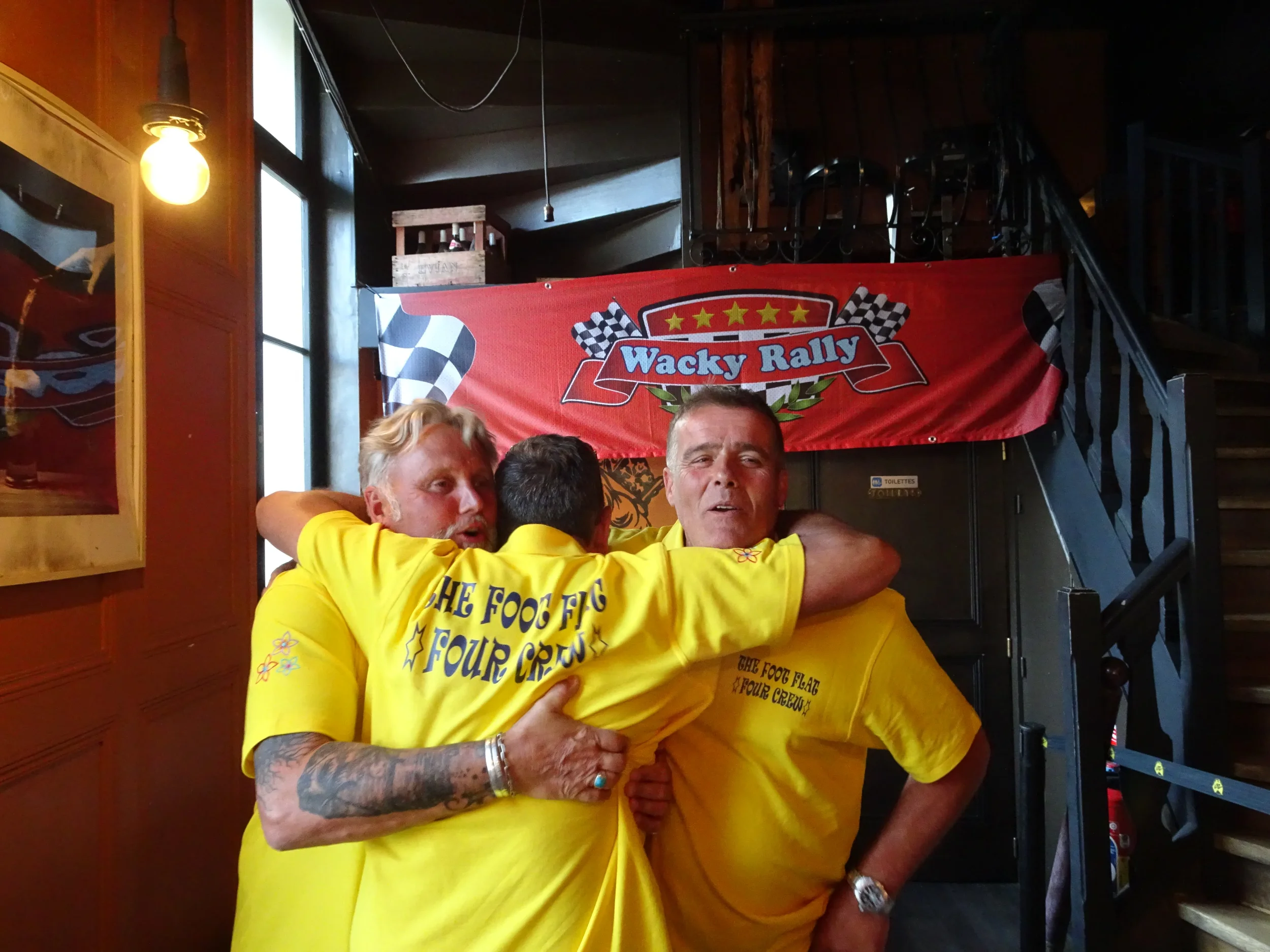 Three men wearing bright yellow shirts embracing each other in a group hug inside a room with wooden walls and a staircase. Behind them is a red banner with the text "Wacky Rally" and checkered flags.