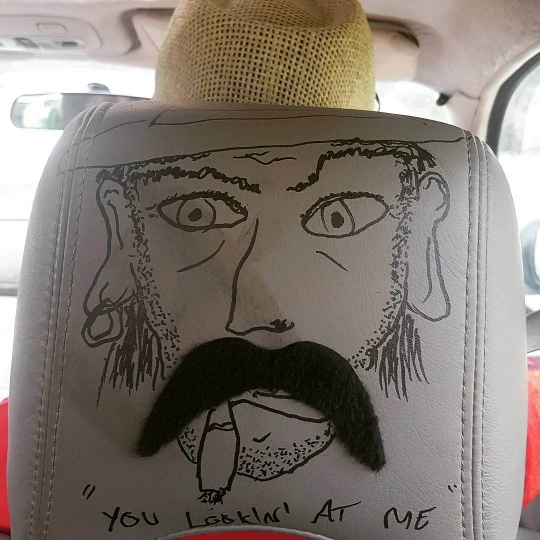 A drawing on a car headrest resembling a man with a mustache and beard, with a piece of black material as the mustache. The drawing includes exaggerated facial features and the phrase "You lookin' at me" written below.