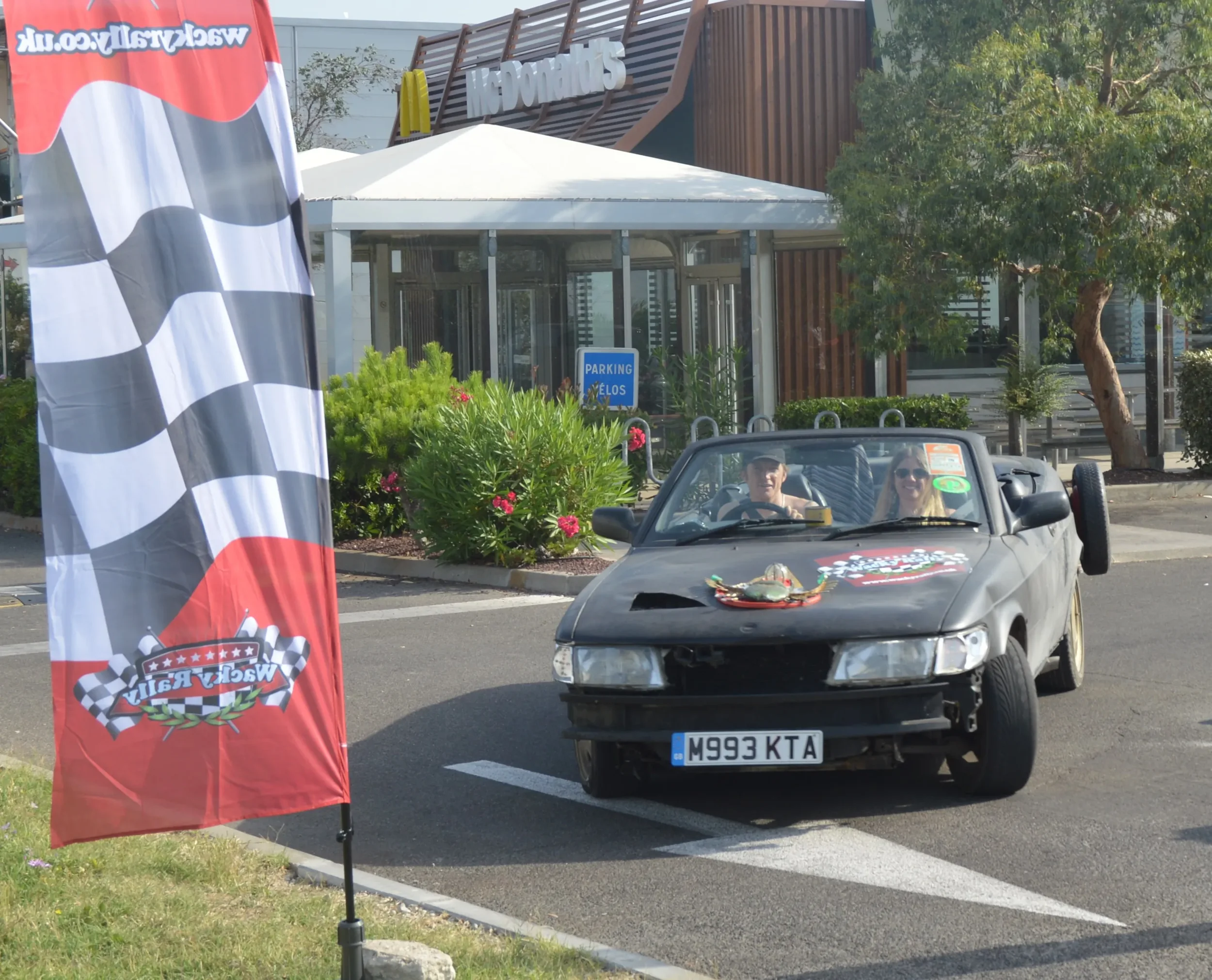 A black convertible car with a missing front grille and a license plate reading M993 KTA, parked in a lot near a building with a sign that says 'Svedarnas'. There are two women inside, smiling, and a flag with a racing checkered pattern and a logo fo