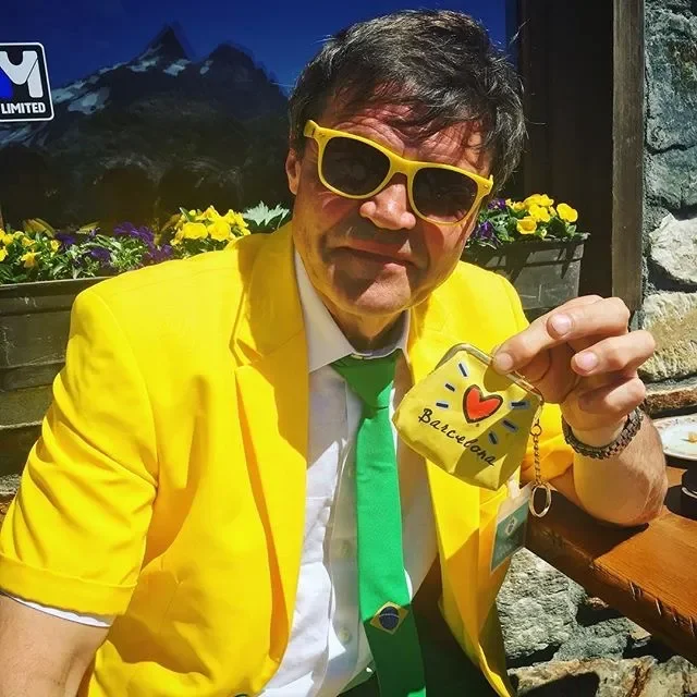 A man dressed in a bright yellow jacket and green tie, wearing yellow sunglasses, holds a small bag with a heart and the word 'Brazil' written on it. He is sitting outdoors with a backdrop of mountains and flowers.