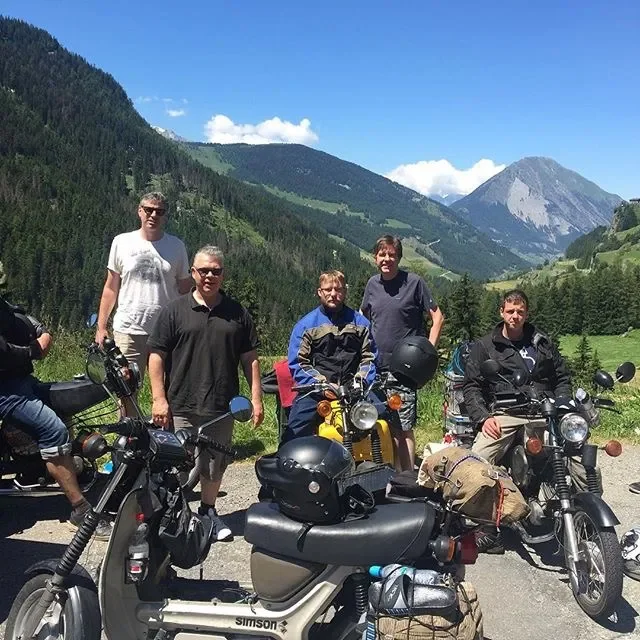 Group of five men standing next to their motorcycles in a mountainous outdoor setting with green hills and a clear blue sky.
