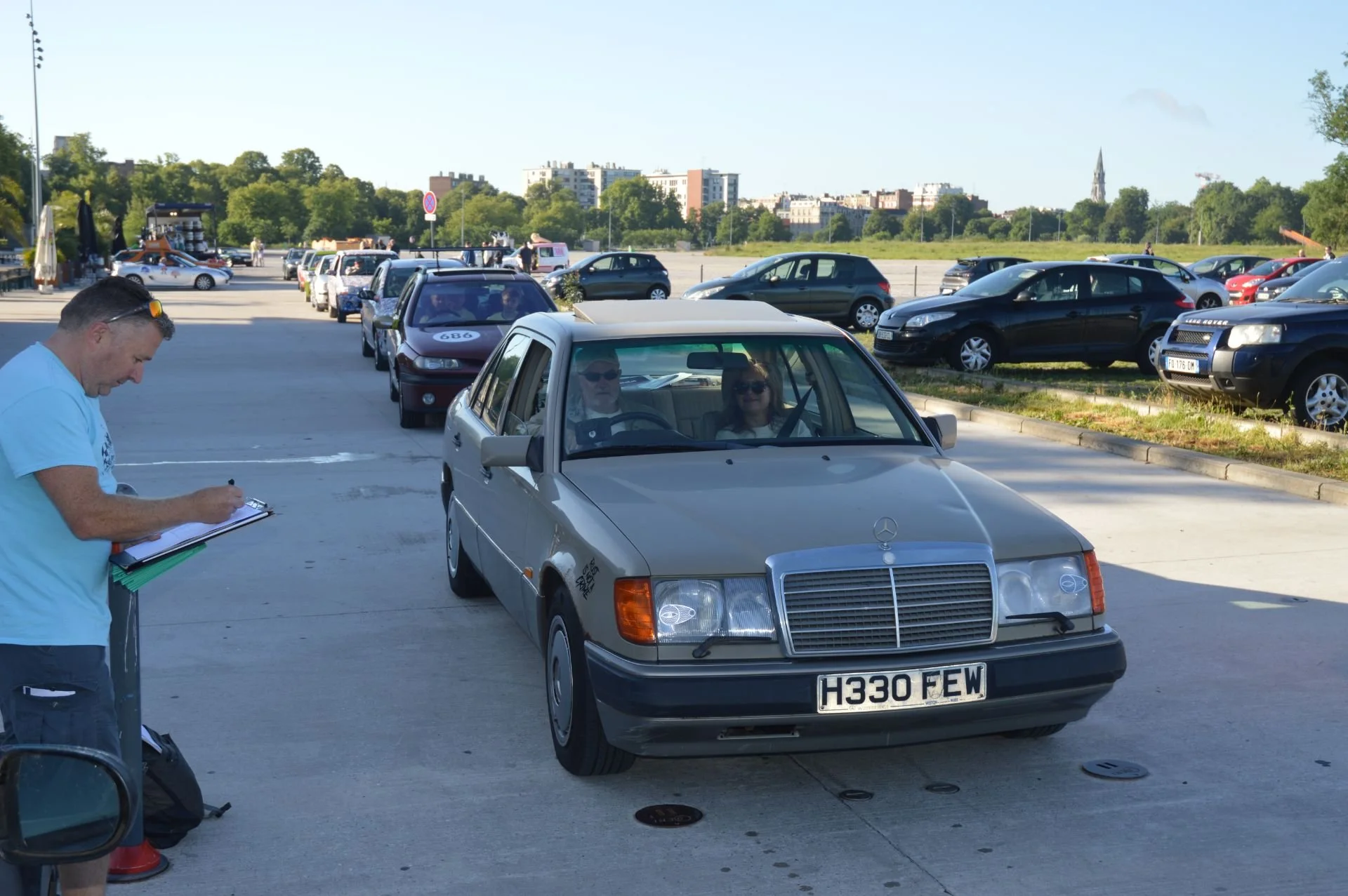 A silver vintage Mercedes-Benz car with a license plate H330 FEW parked in a busy parking lot on a sunny day with multiple other cars in the background.