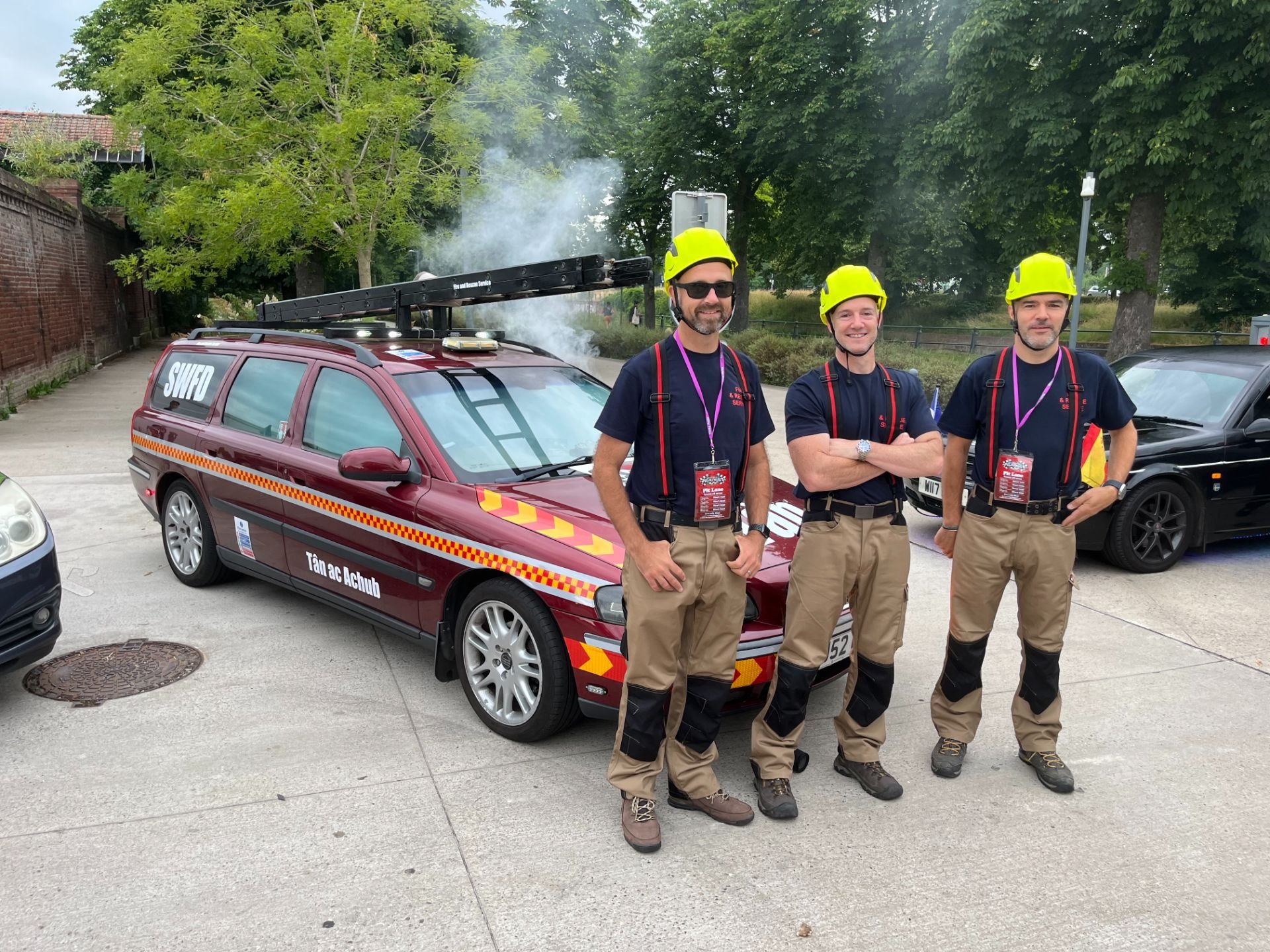 Three firefighters in yellow helmets standing in front of a red emergency response vehicle with text in German and an open ladder on top, on a paved area with trees in the background.