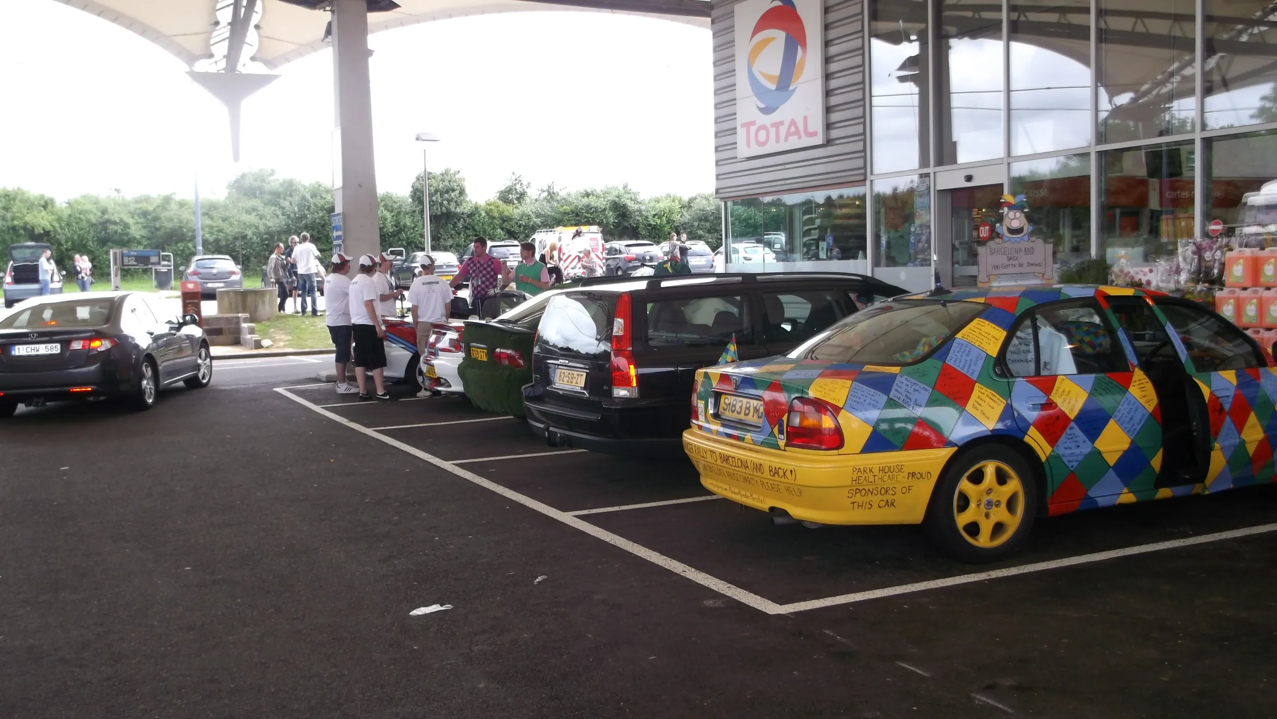 A parking lot outside a building with several cars parked, including a colorful car with a yellow bottom and a pattern of colored squares. People are standing near cars and entering the building, which has a large sign that says 'TOTAL' and a cartoon