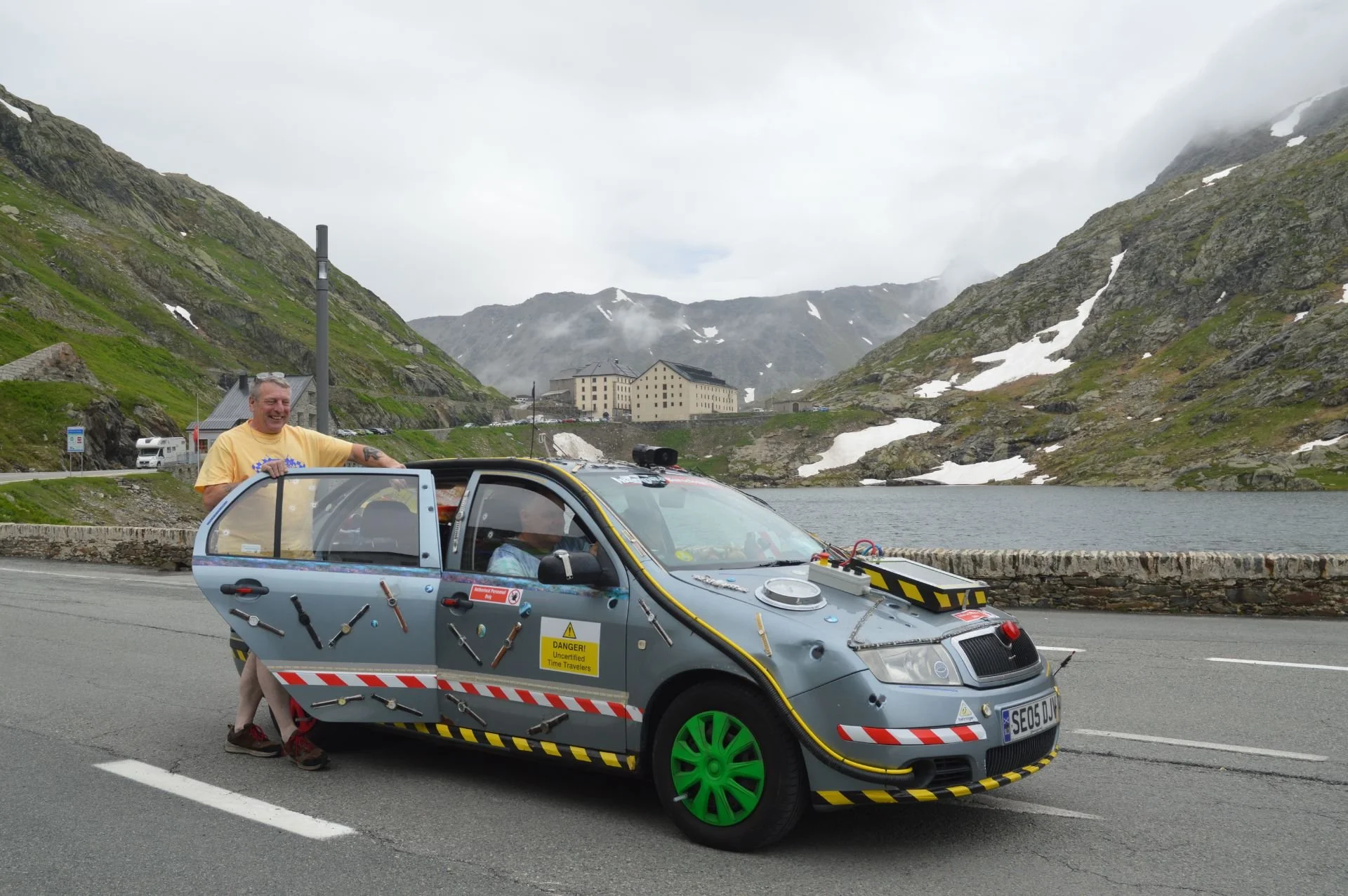 A man standing next to a small experimental car with various sensors and modifications, parked by a lake with mountains and cloudy sky in the background.