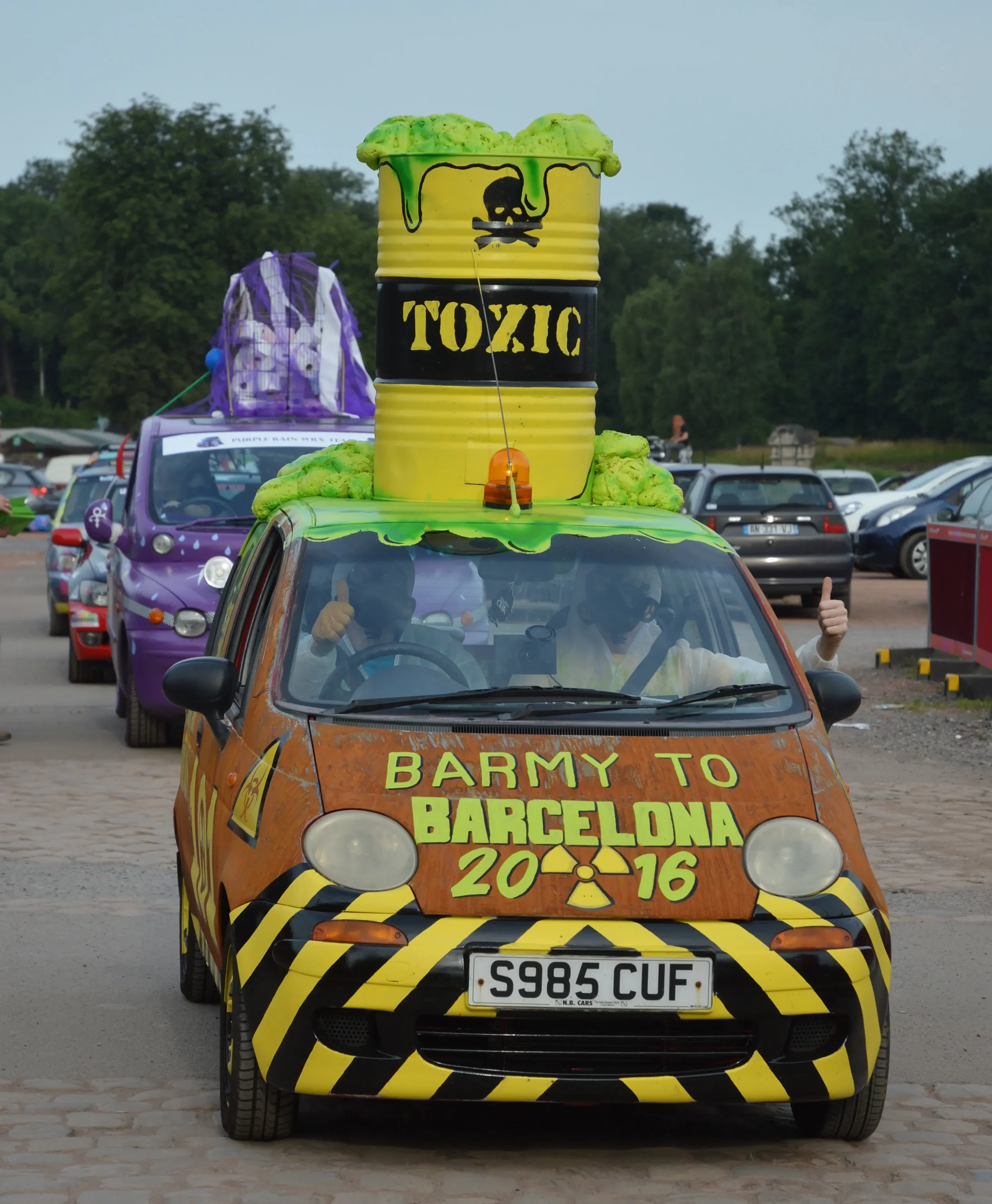 A small, colorful car decorated with warning stripes and a warning sign, carrying a large yellow barrel labeled toxic on its roof, with two people inside giving thumbs up in a parking lot.
