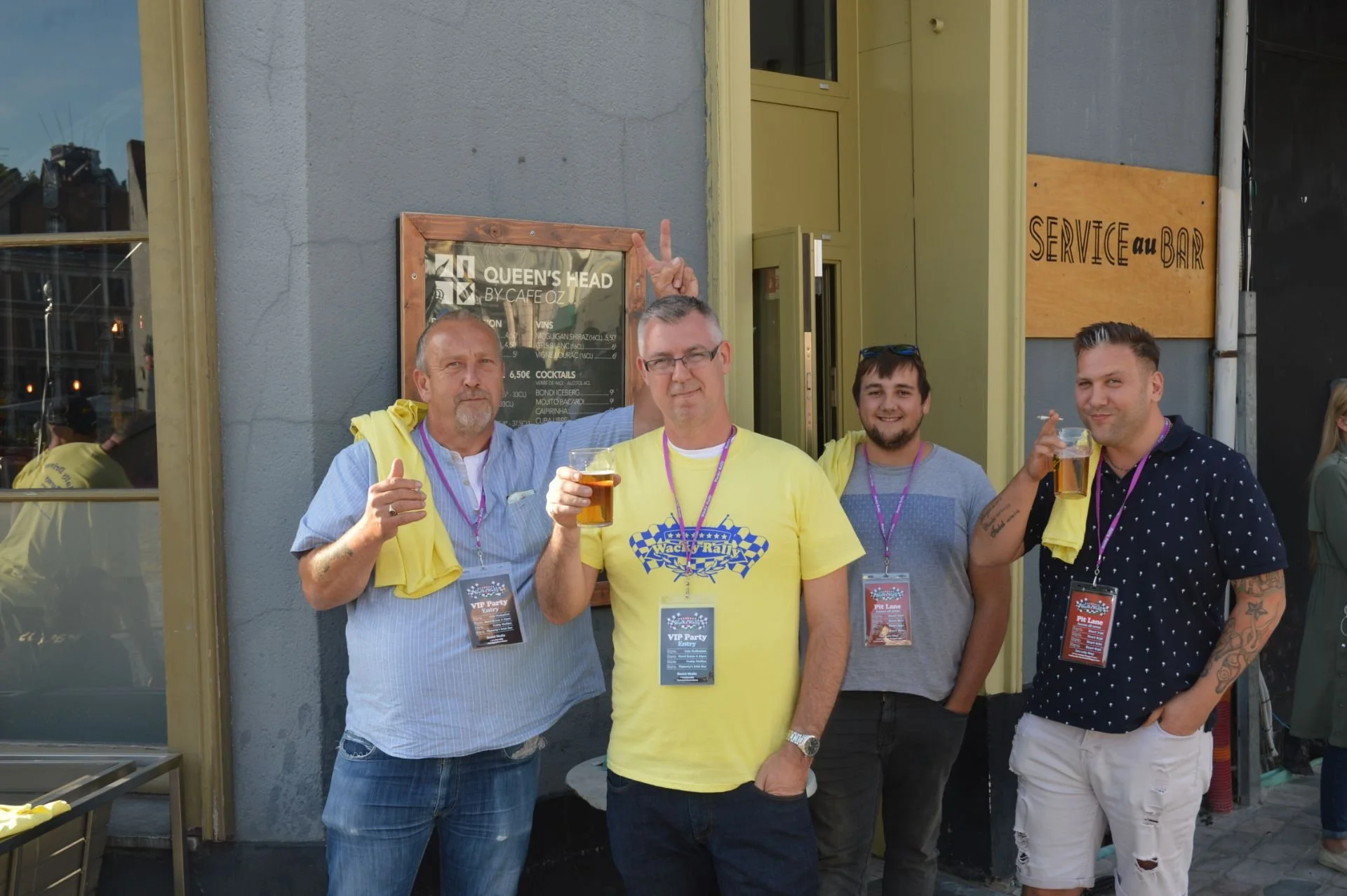 Four men standing outside a bar drinking beer, two of them are holding glasses of beer and wearing VIP party badges, one man is making a peace sign with his fingers behind a man's head.