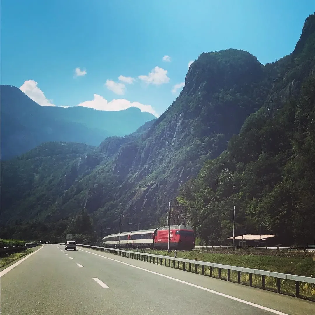 A highway running through a mountainous landscape with a red and white train passing by on the side, under a bright blue sky with scattered clouds.