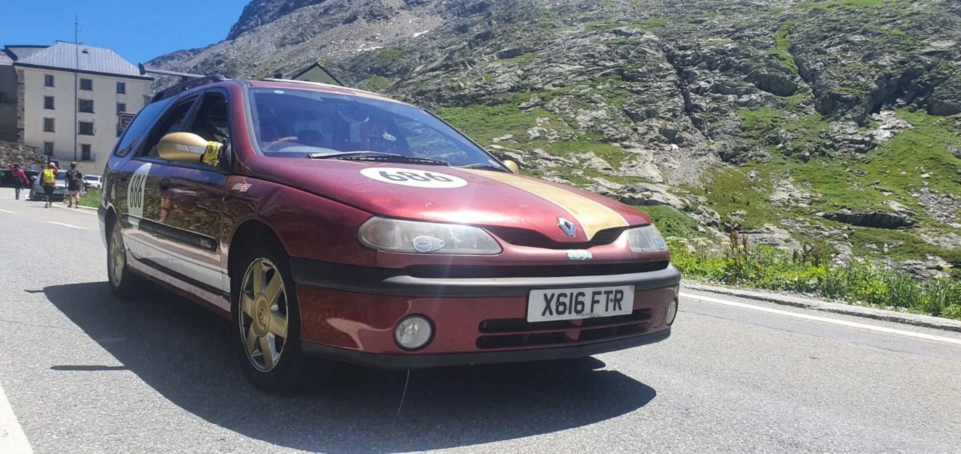 A maroon Renault car with gold accents and wheels, parked on a mountain road during a race event with the number 696 on the hood and a Scottish license plate X616 FTR. There are people and buildings in the background with rocky mountain terrain and g