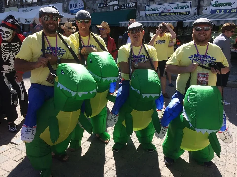 Four men wearing yellow shirts, blue pants, and inflatable dinosaur costumes on their laps, standing outdoors at a street event with other people around.