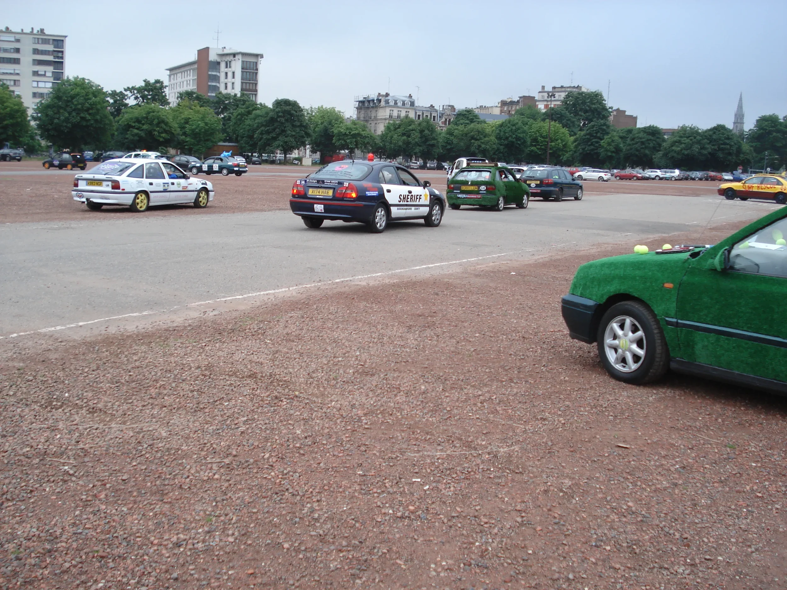 Police cars and a sheriff vehicle in a parking lot with trees and buildings in the background.