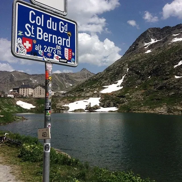 Signpost with the text 'Col du Gd St Bernard' and an altitude of 2,473 meters, next to a mountain lake with snow patches and mountain buildings in the background.