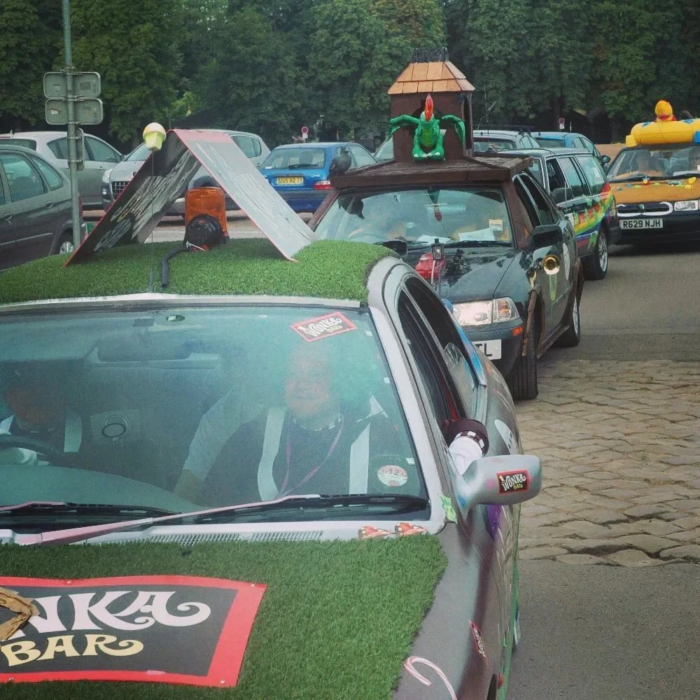 Line of cars decorated for a parade, including a car with a green dragon and castle on top, a car with a grass-covered hood, and a hot dog float in the background.