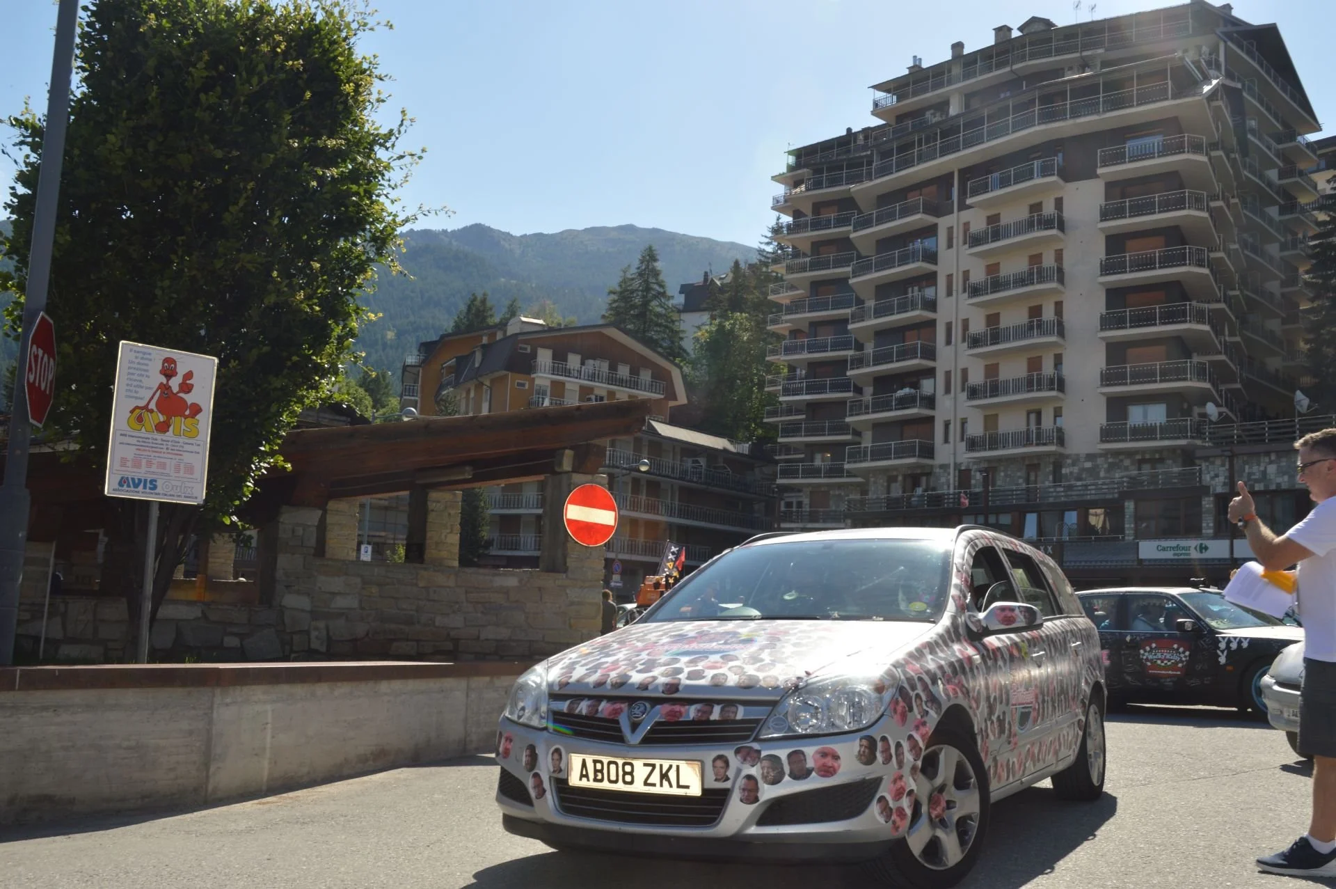 A decorated silver car with face cutouts covers the hood and bumper, driving past a man with sunglasses and a white shirt holding papers. The background shows tall buildings, trees, and mountain hills under a clear blue sky.