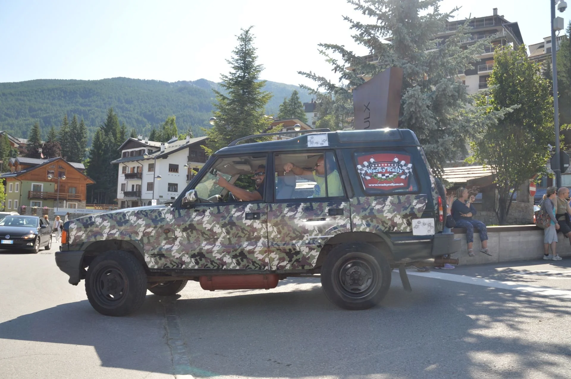 A vehicle with a camouflage pattern parked on a street with mountains and residential buildings in the background. There are people sitting on a ledge and standing nearby. Two men inside the vehicle are interacting, one reaching out to the other.