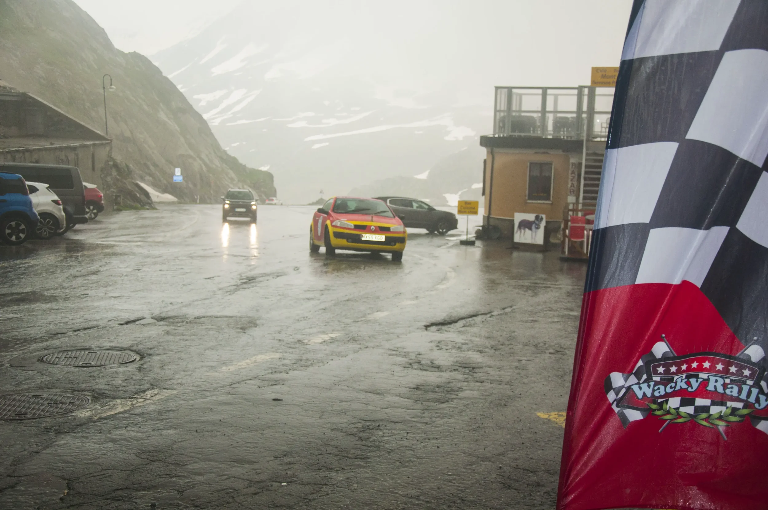 Rainy mountain roadside with parked cars, a yellow and red race car, and a checkered flag banner reading 'Wacky Rally'.