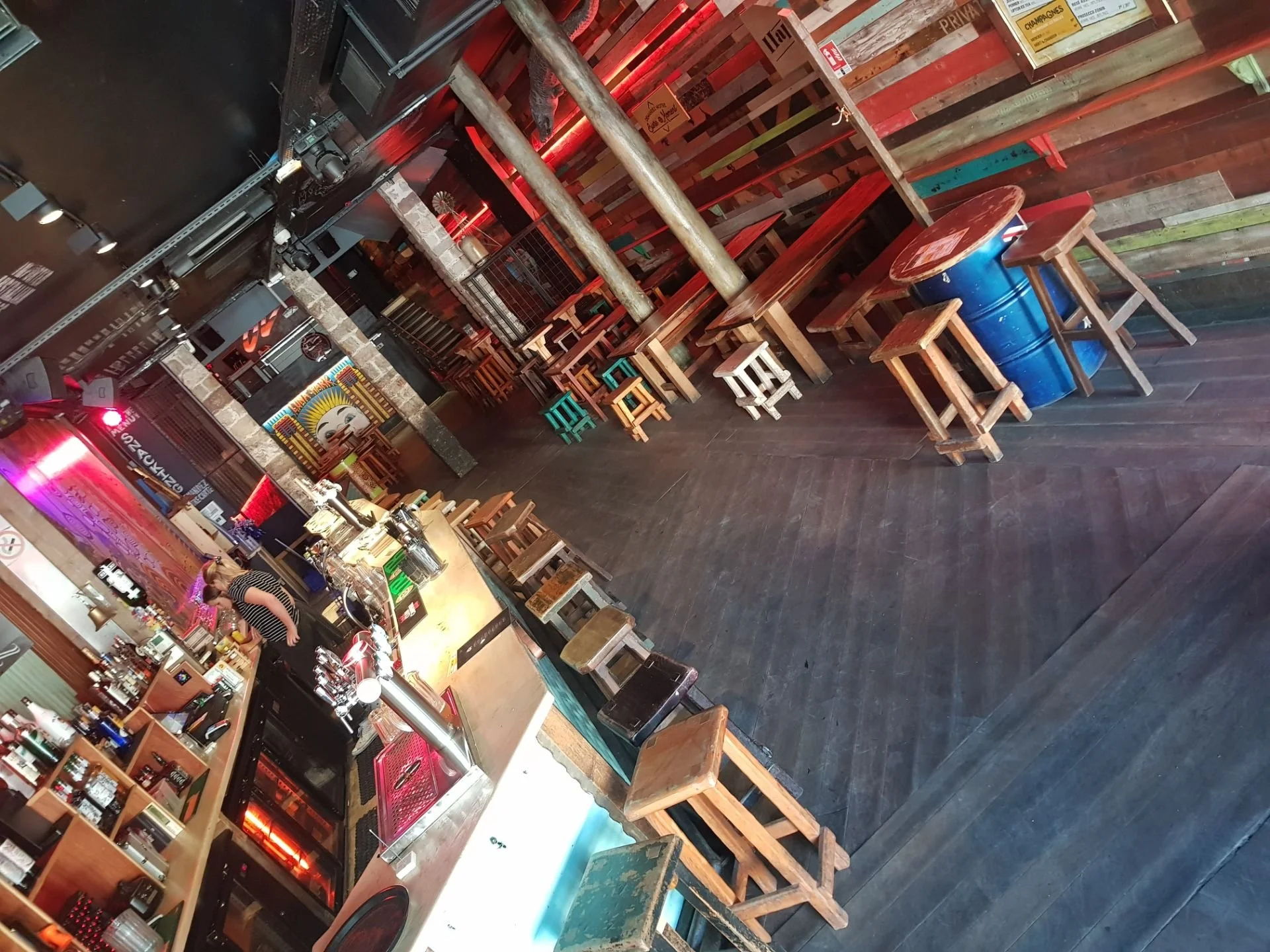 Interior of a bar or restaurant with a wooden counter, colorful stools, high tables, and a rustic wooden wall with artwork and decorations.