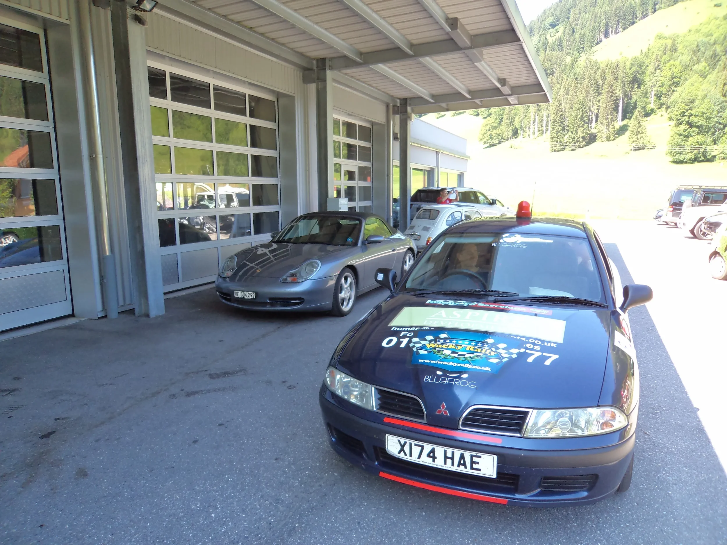A row of parked cars outside a building with large garage-style doors, including a black Mitsubishi with rally stickers and a silver Porsche convertible, with a mountainous green landscape in the background.