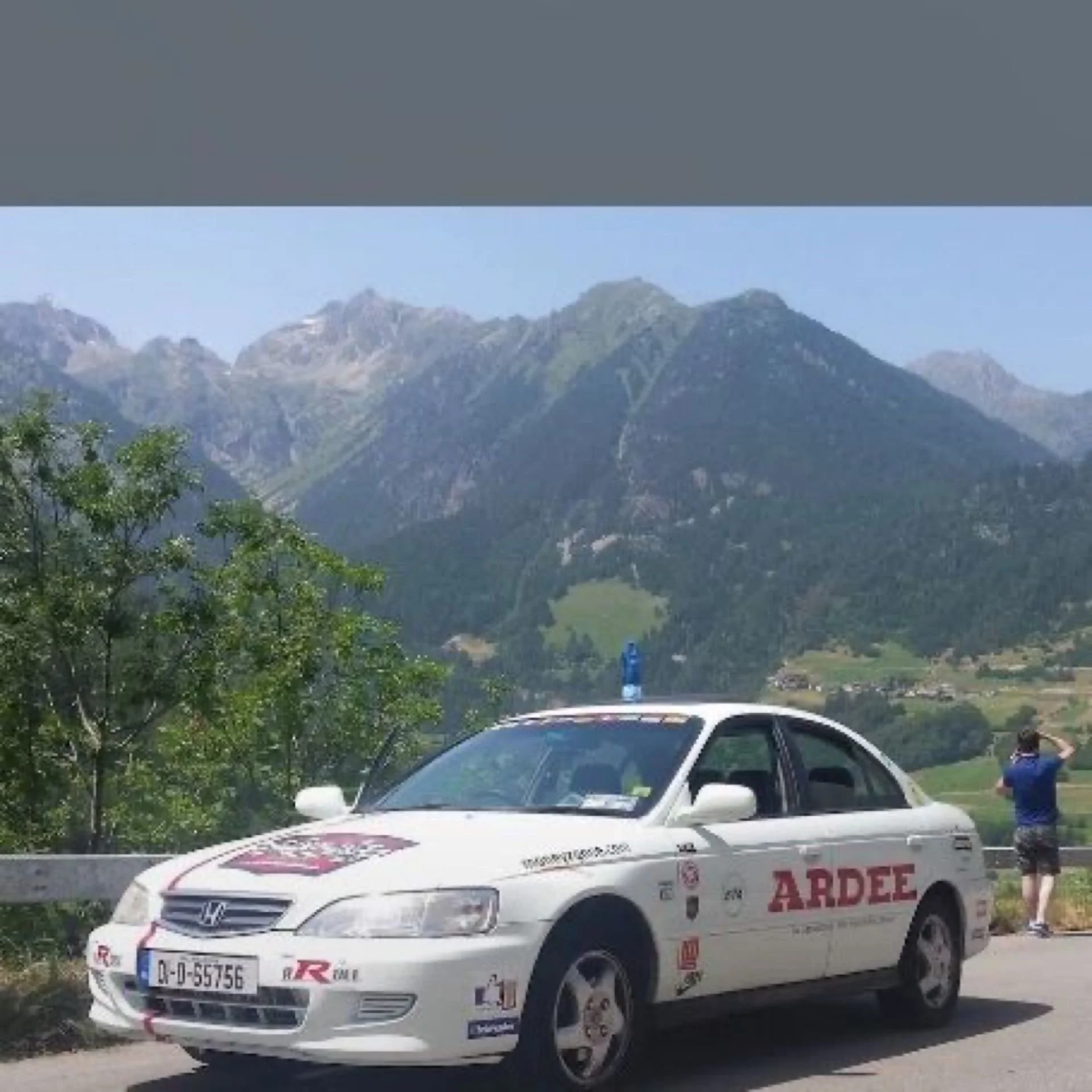 A white Honda car with ARDEE branding and various stickers on it, parked alongside a mountain road with green trees, mountain peaks, and a person in blue taking a photo in the background.