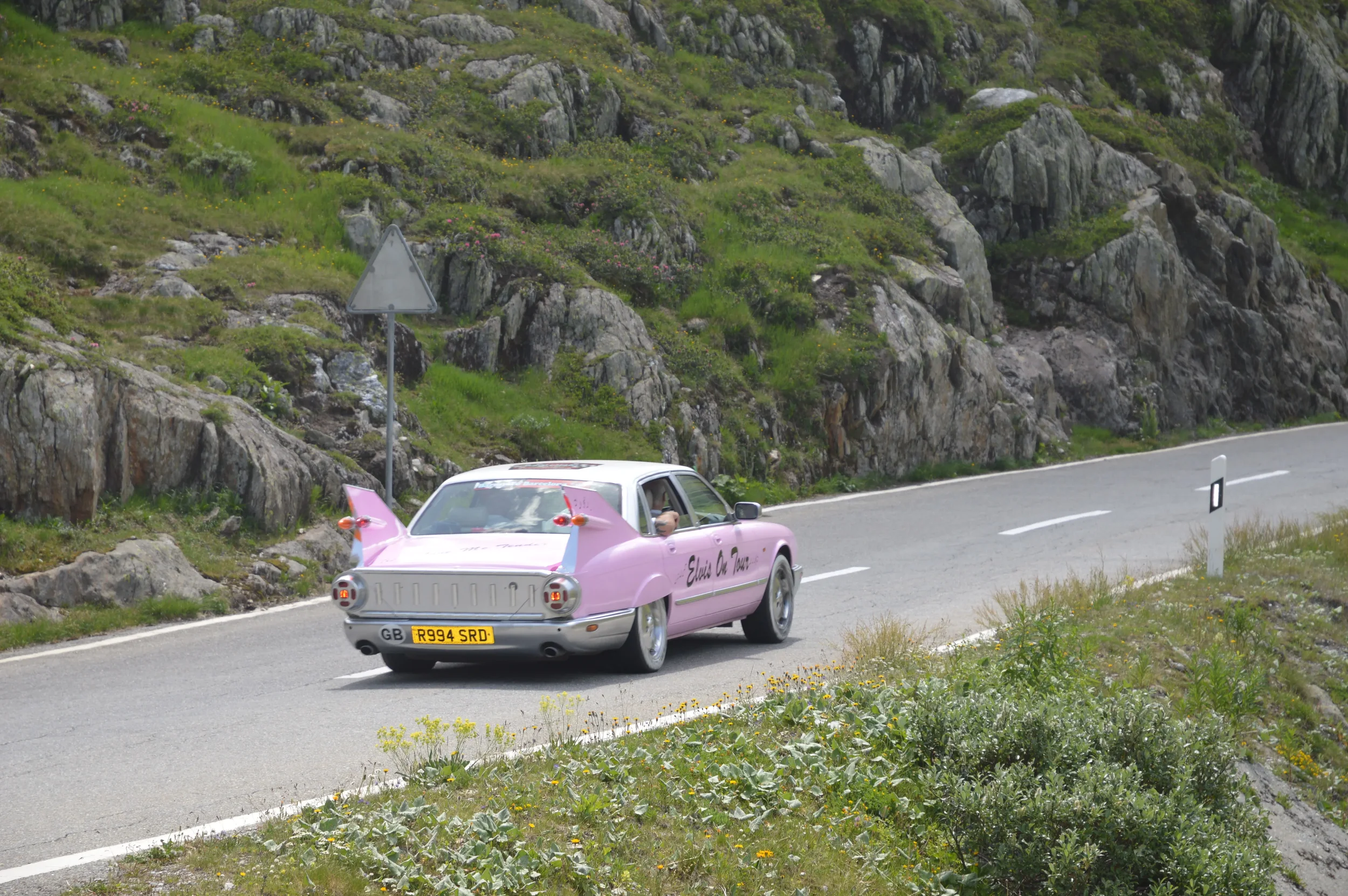 A pink vintage car with costume wings driving on a mountain road, with rocky terrain and green vegetation in the background.