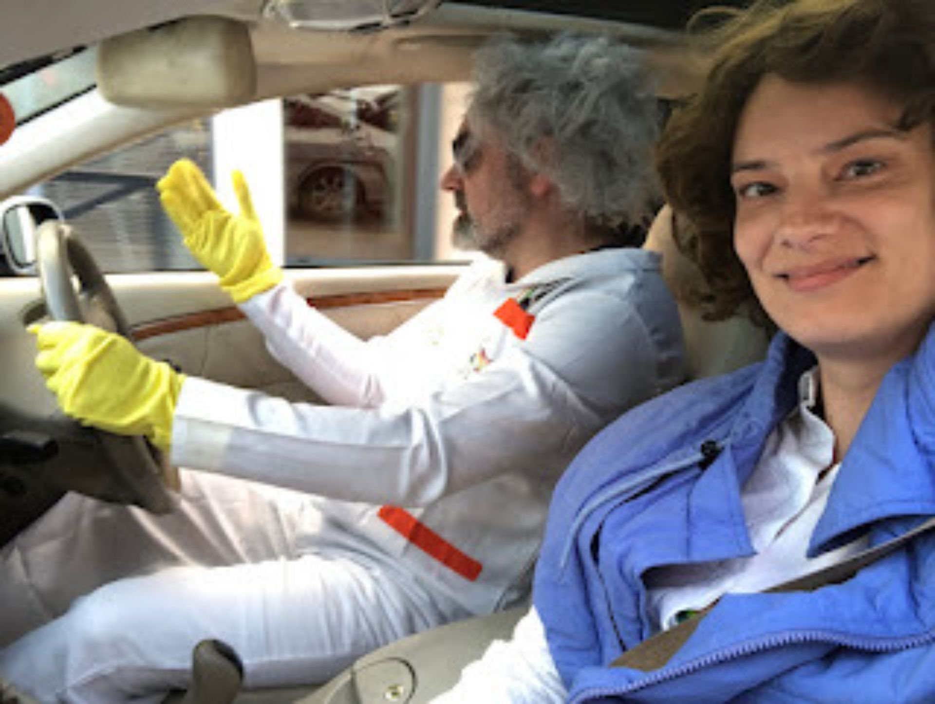 Two people sitting in a car, one in the driver's seat and the other in the passenger seat. The driver is wearing a white suit with yellow gloves, and is gesturing with their hand. The passenger is smiling, wearing a blue jacket and has brown hair.