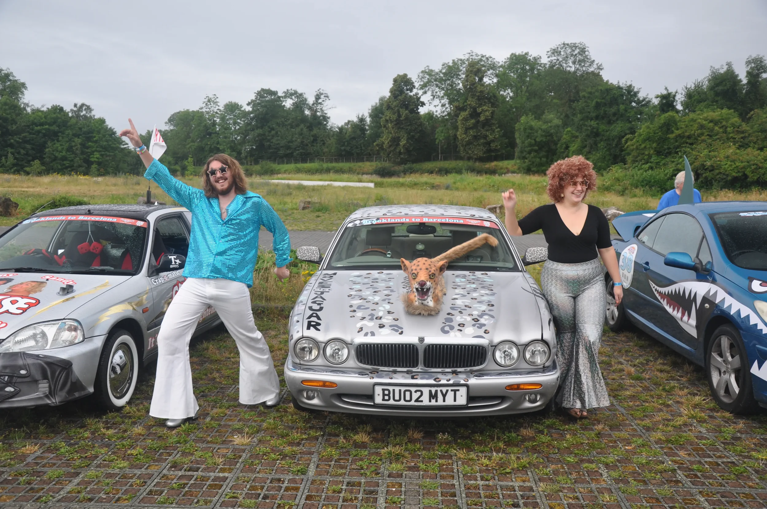 Two joyful people posing next to a silver car with a fake lion on the hood at an outdoor car show, flanked by other creatively decorated cars.