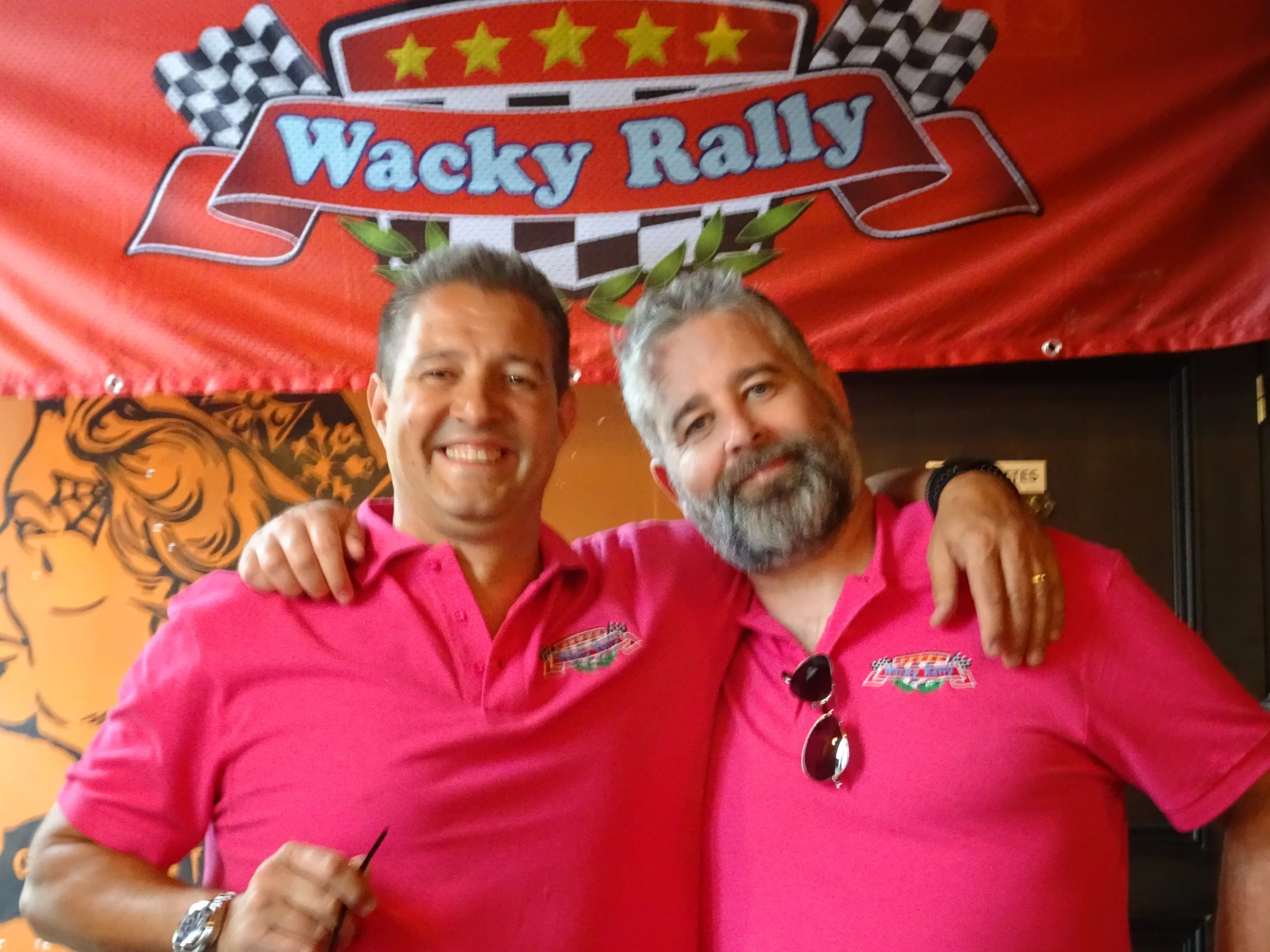 Two men wearing matching pink shirts, smiling and arm-in-arm, with a red banner that reads 'Wacky Rally' and two checkered flags in the background.