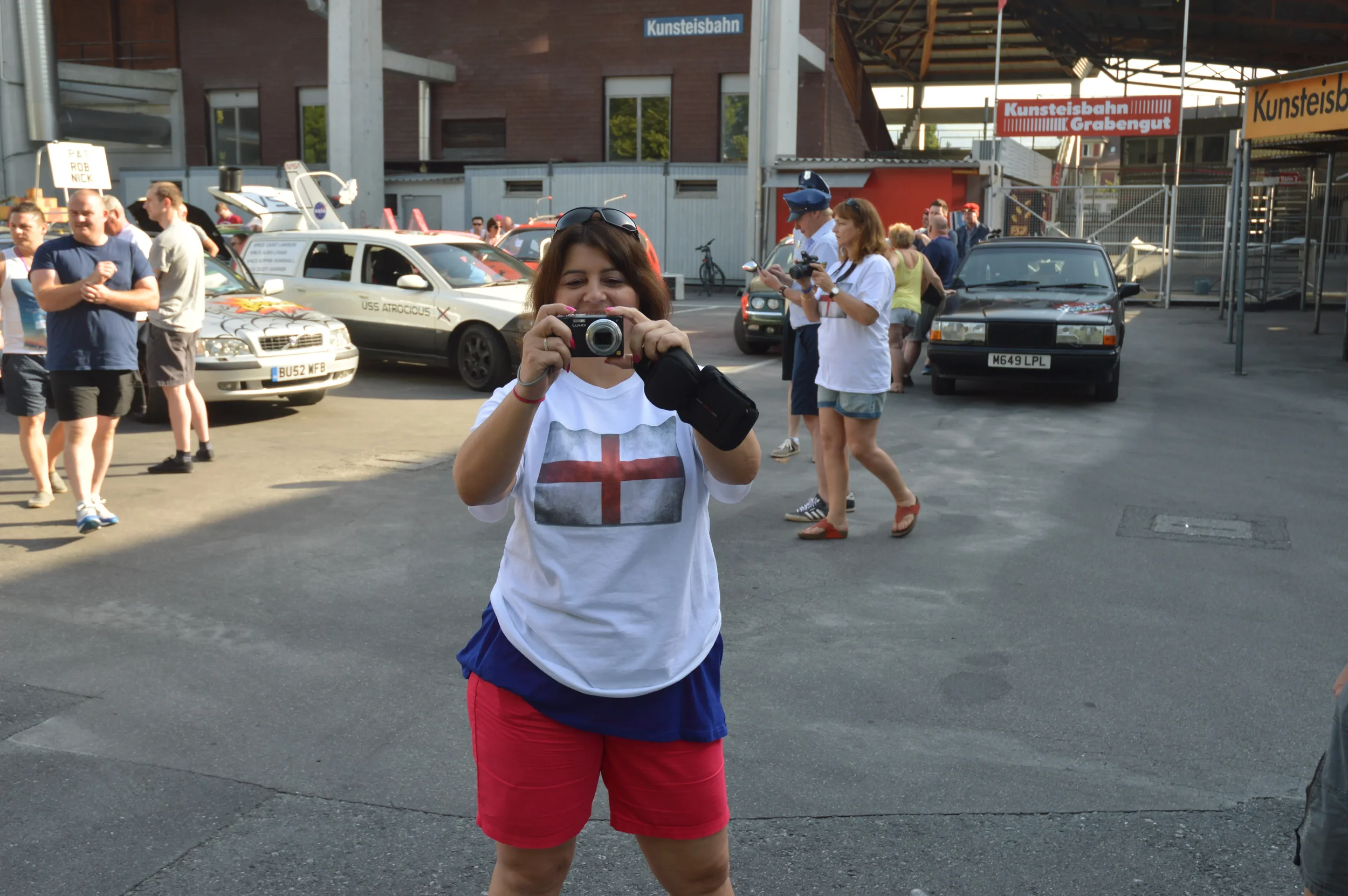 A woman taking a photo with a camera at a gathering near cars and bystanders, with signs indicating an event at a train station.