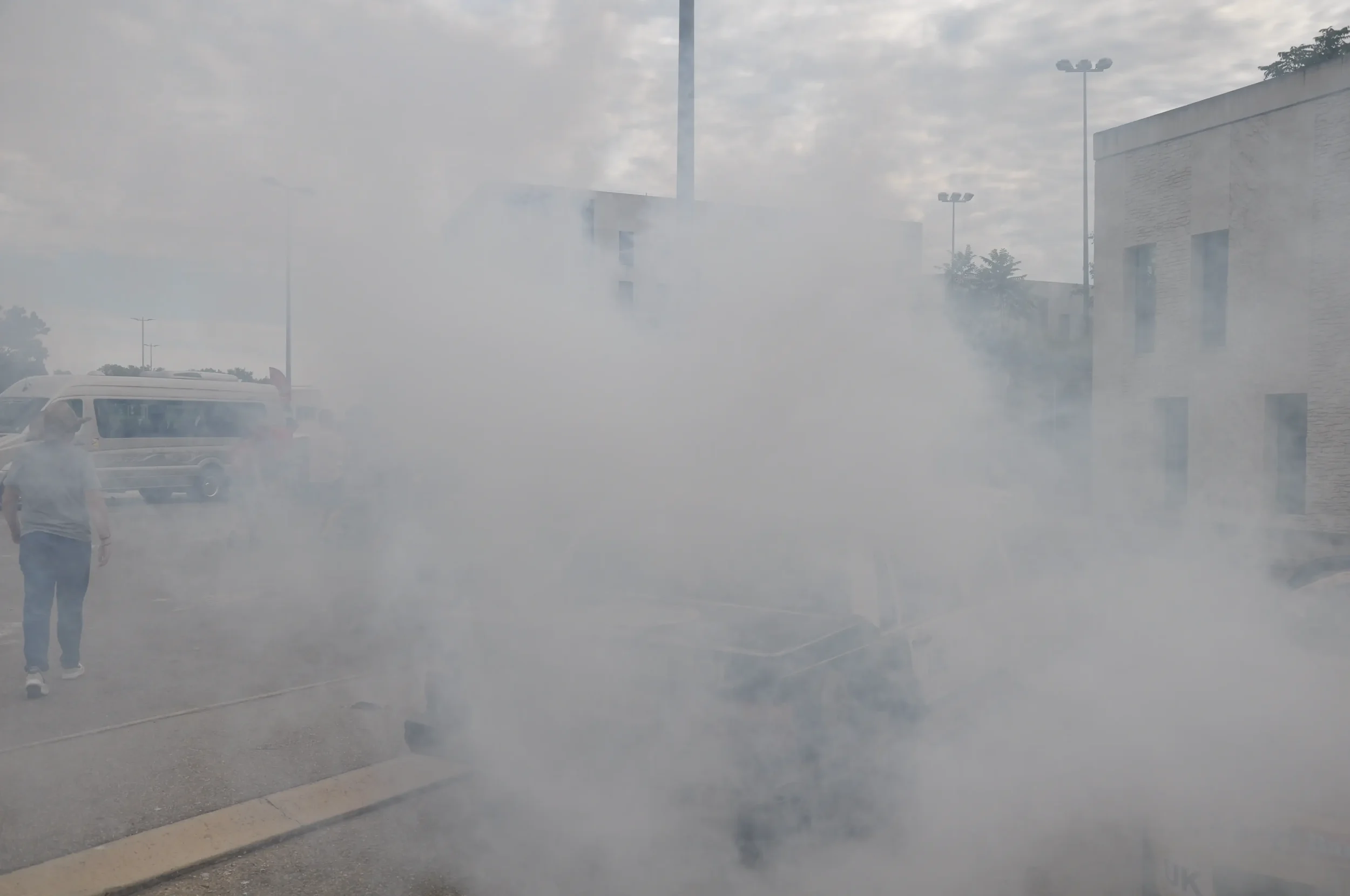 A person standing near a cloud of smoke or fog in an outdoor parking lot with buildings and vehicles in the background.