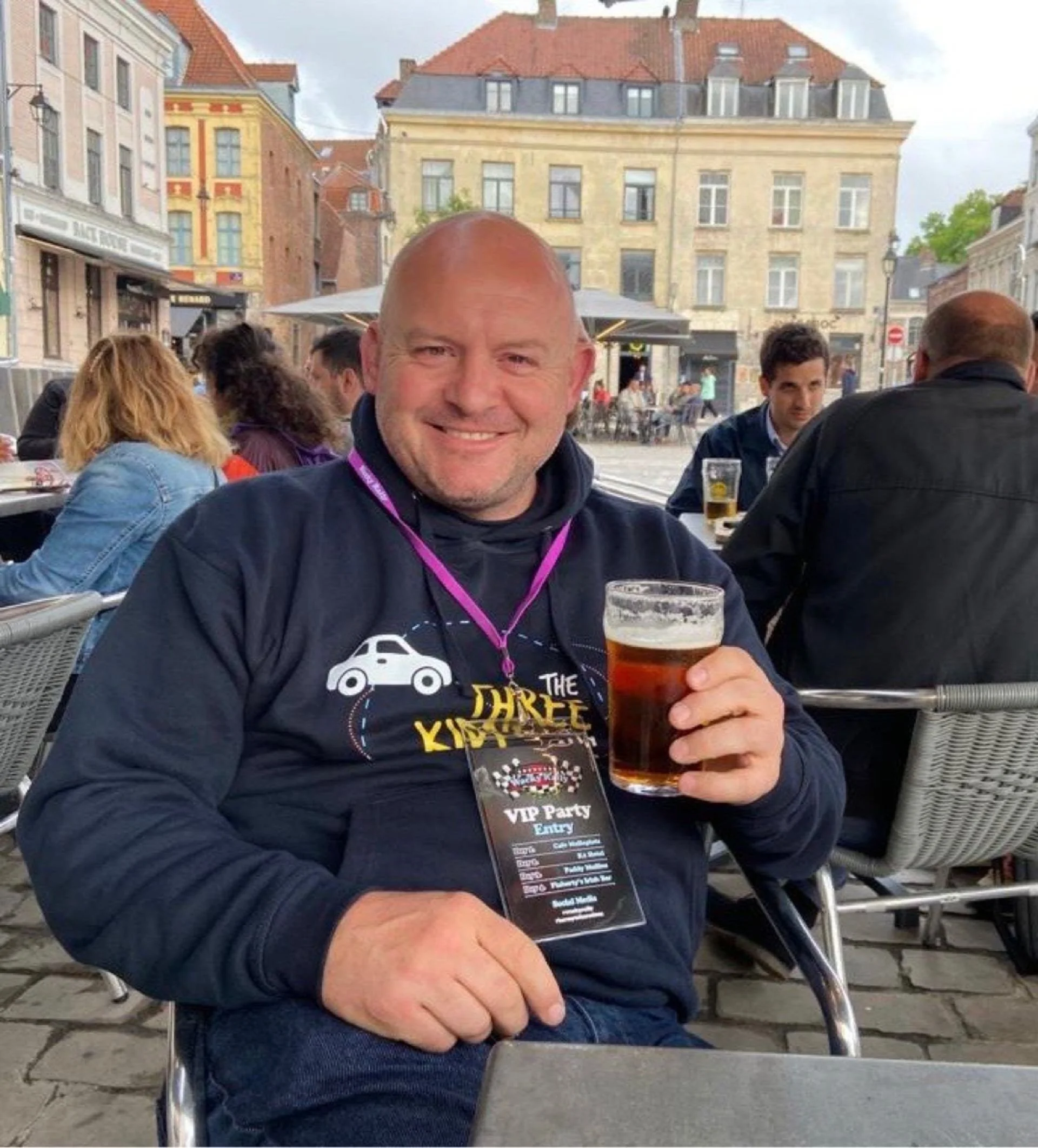 A smiling man holding a pint of beer sitting at an outdoor cafe, wearing a black hoodie with a graphic and a VIP badge around his neck, with other people and buildings in the background.