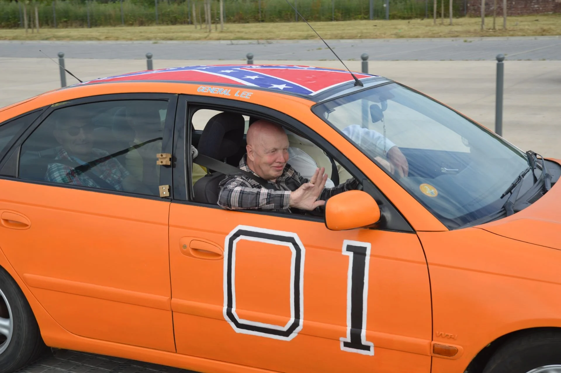 A man with a shaved head sitting in an orange race car with the number 01 painted on the side, holding up his hand in a stop gesture, with a passenger in the back seat and another person partially visible, outside on a paved lot.