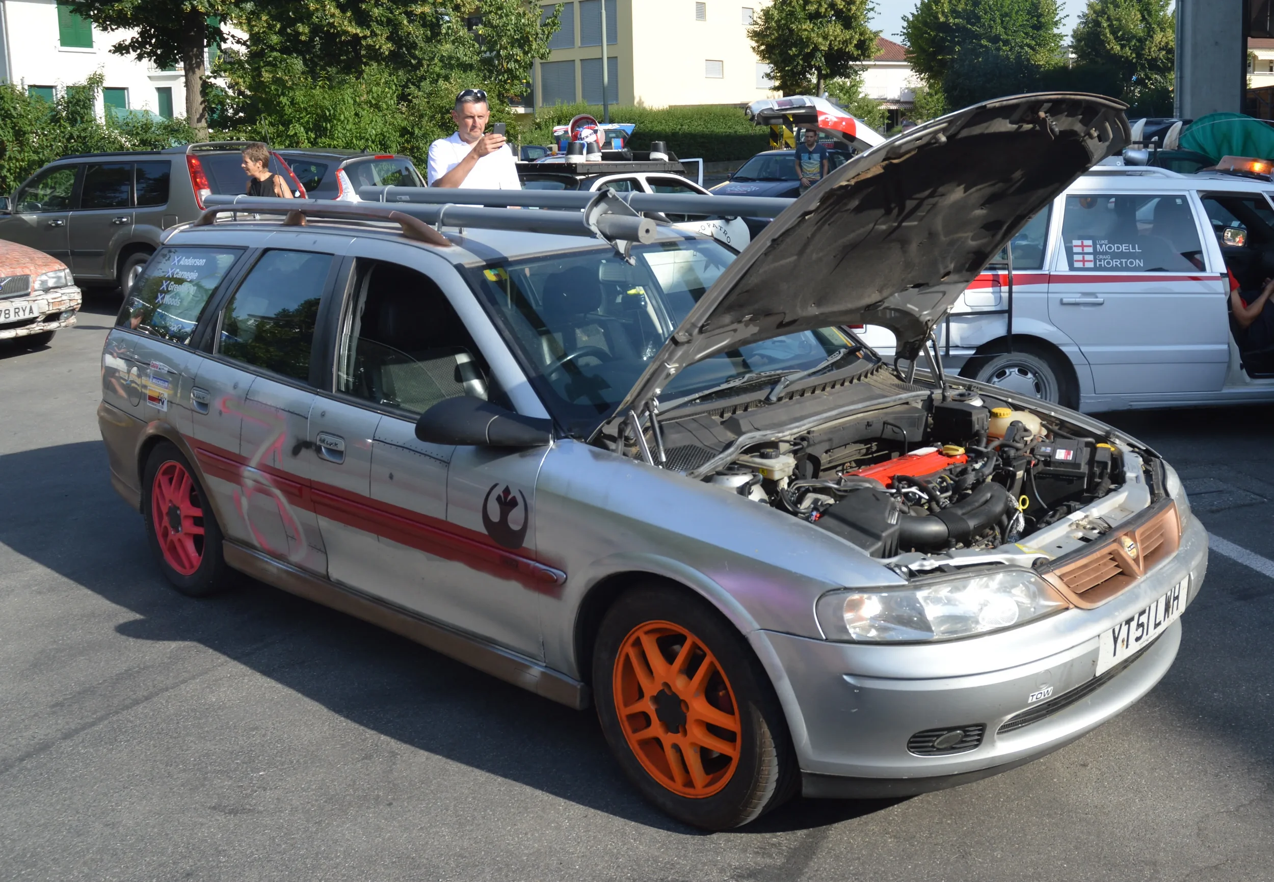 A silver station wagon with a Star Wars Rebel Alliance logo on the side, colorful wheels, and an open hood displaying its engine, parked in a lot with other vehicles and people around.