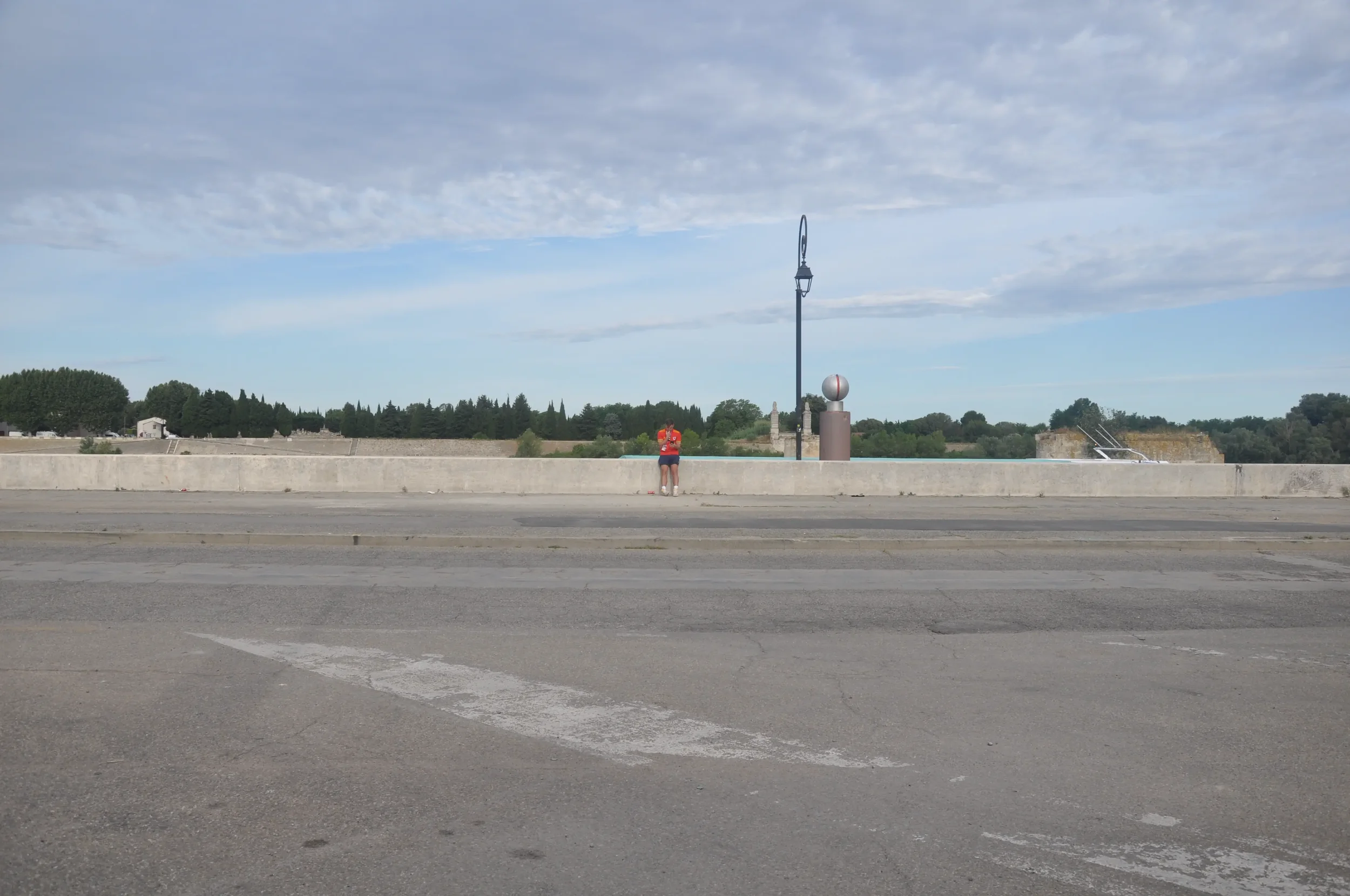 A person standing on a sidewalk near a lamppost, with a blue sky and some clouds overhead, and a stone wall in the background.