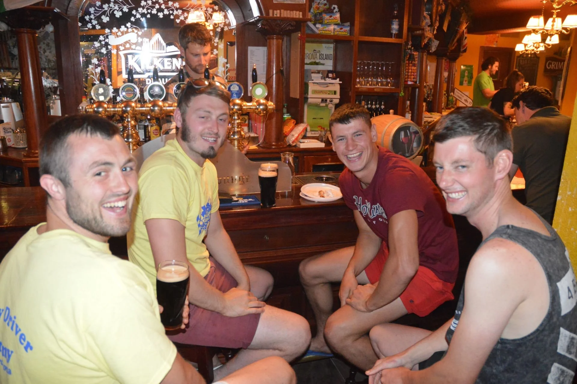 Four young men sitting together at a bar, smiling and laughing. They have drinks in front of them, and the bar has multiple taps and a mirror with the word 'Kiken' reflected. The background shows other patrons and bar shelves with bottles.