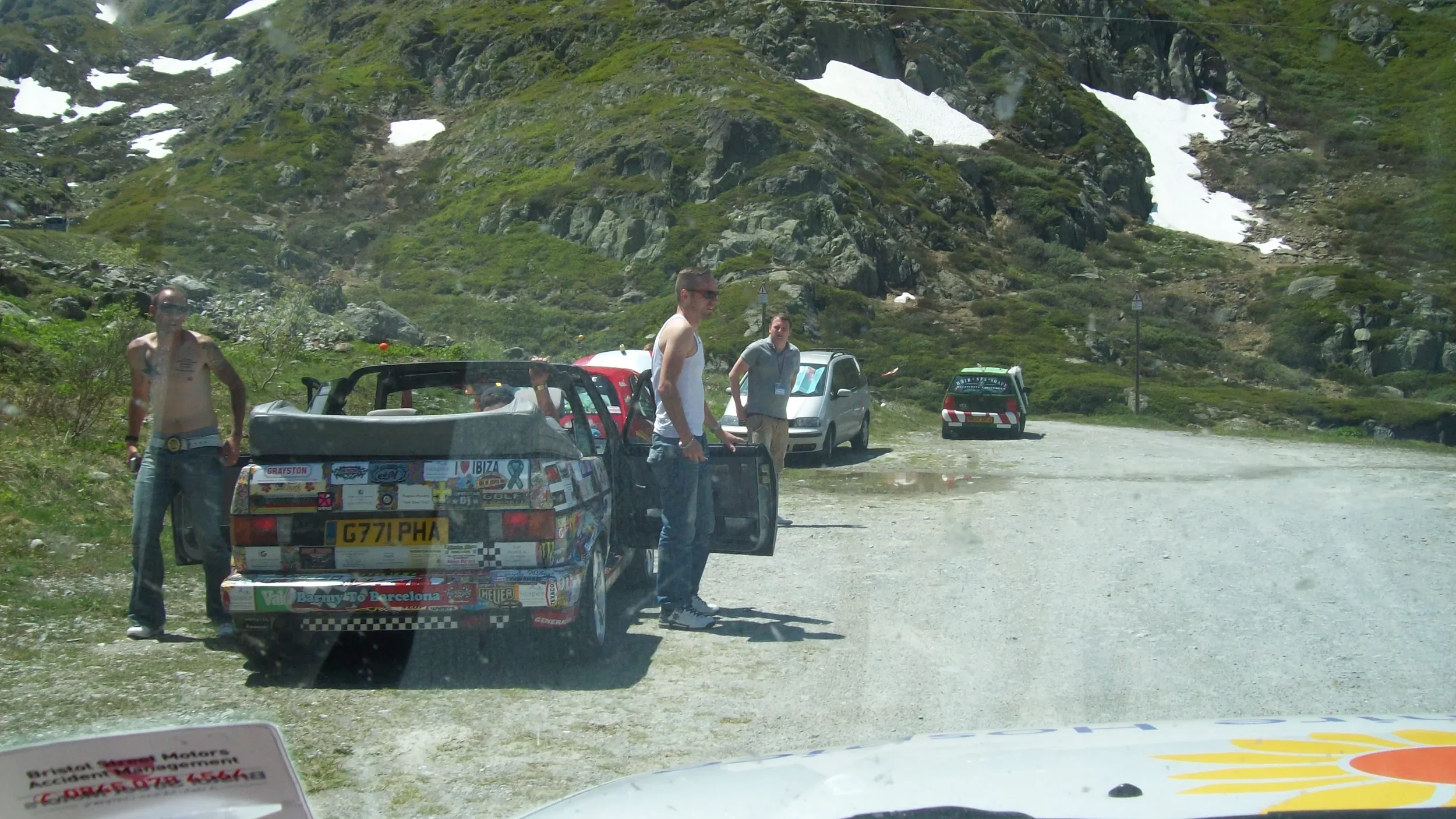 Four men standing outside on a gravel area in a mountainous landscape with patches of snow. Two cars are parked nearby, one with various stickers.