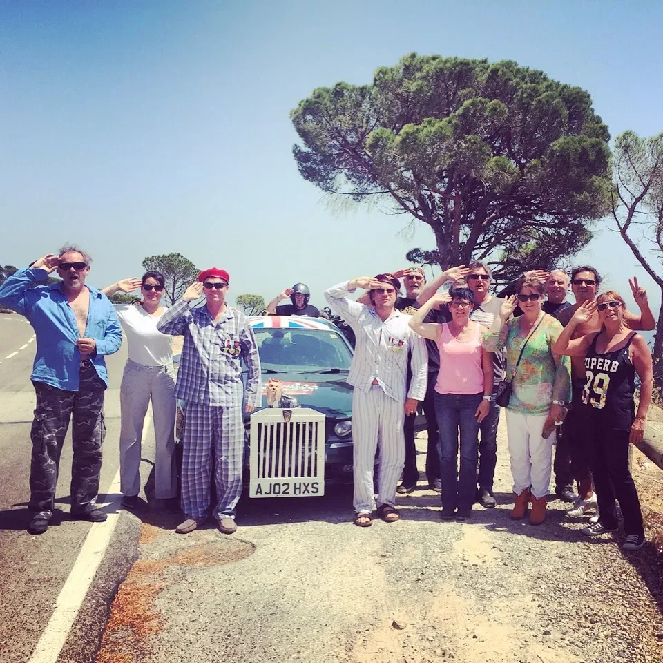 Group of people standing on the side of a road, saluting, with a black car behind them and trees in the background.