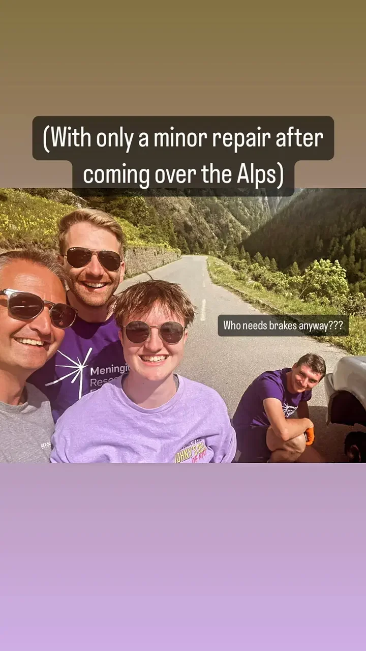 Four people smiling in front of a mountain landscape on a road, with one person sitting on the ground and a car nearby