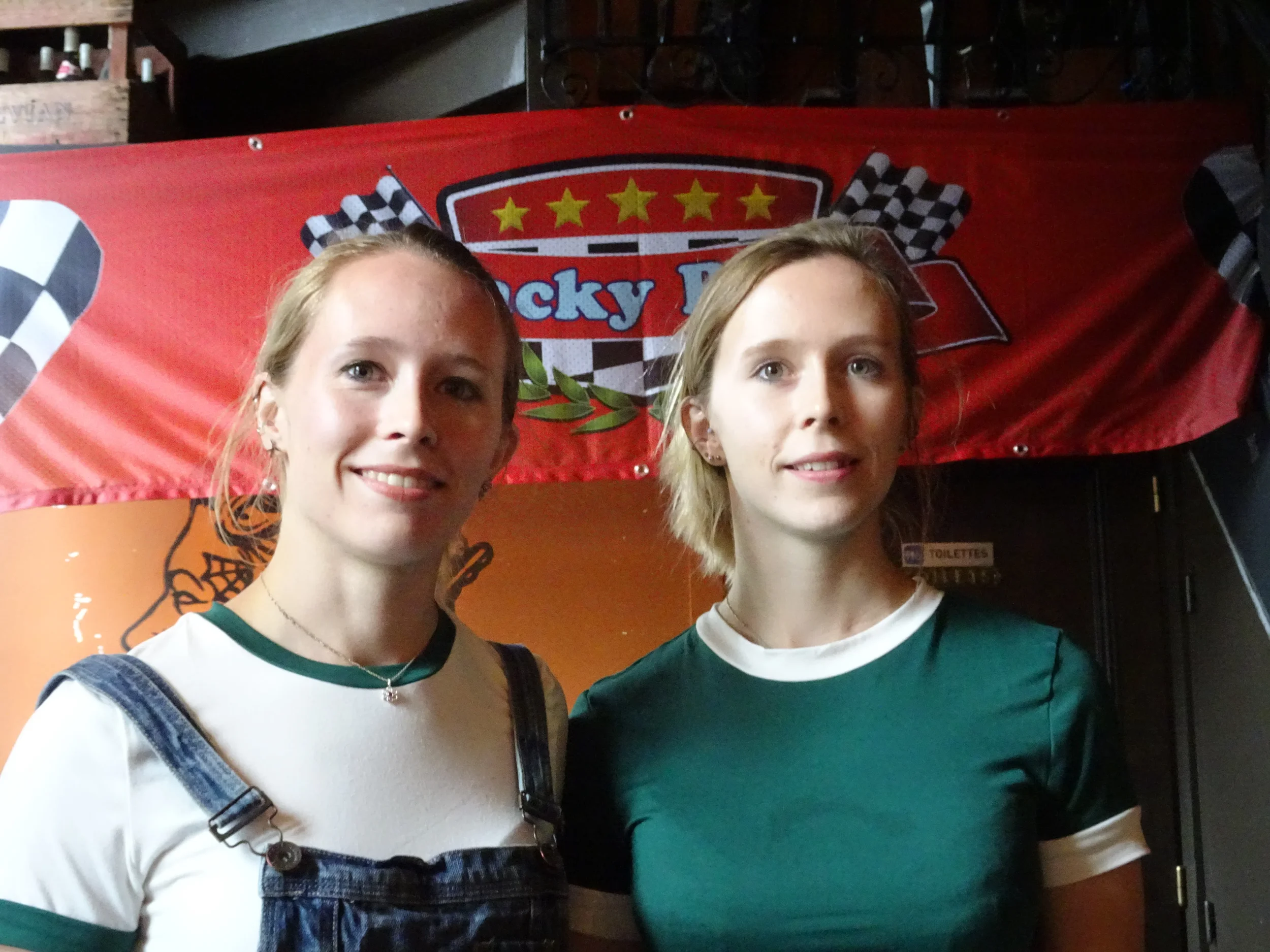 Two young women standing side by side, smiling, in front of a red checkered flag with the words "Lucky D" on it.