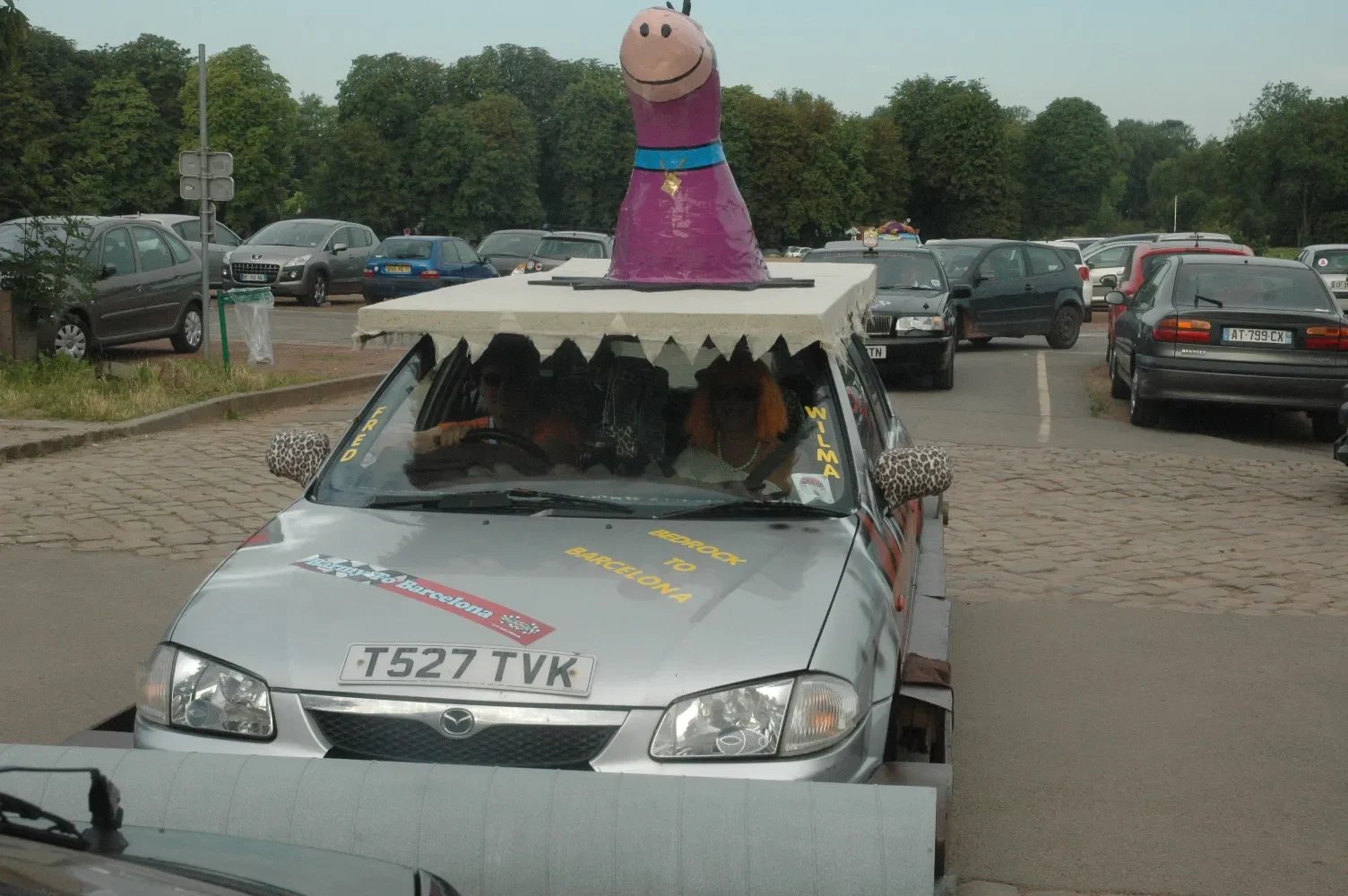 Custom decorated car with a large Peppa Pig figure on the roof, cars in a parking lot, and people inside the car.