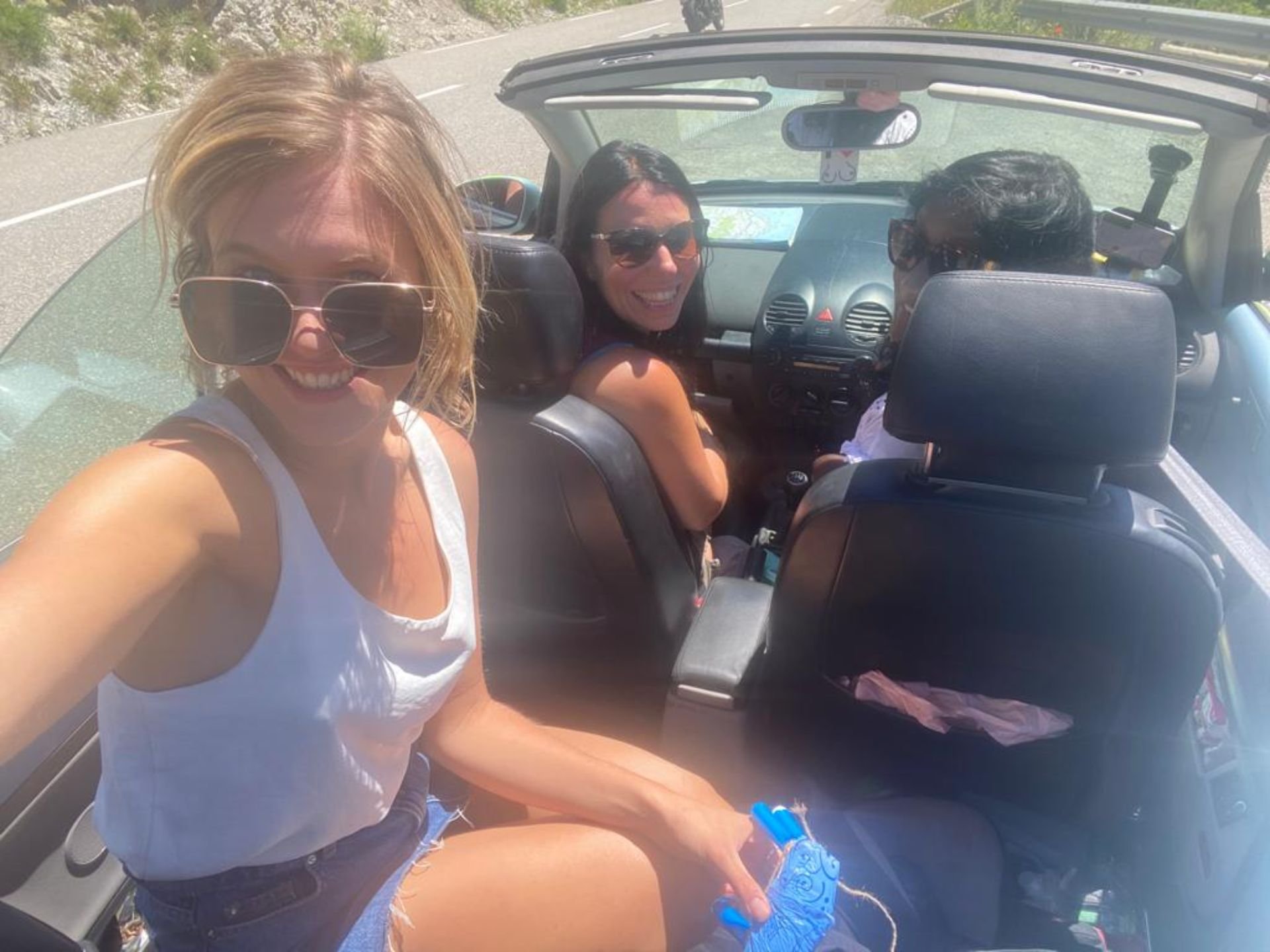 Three women in a convertible car on a sunny day. One woman is taking a selfie, and two women are sitting in the back seat, smiling. All are wearing sunglasses.