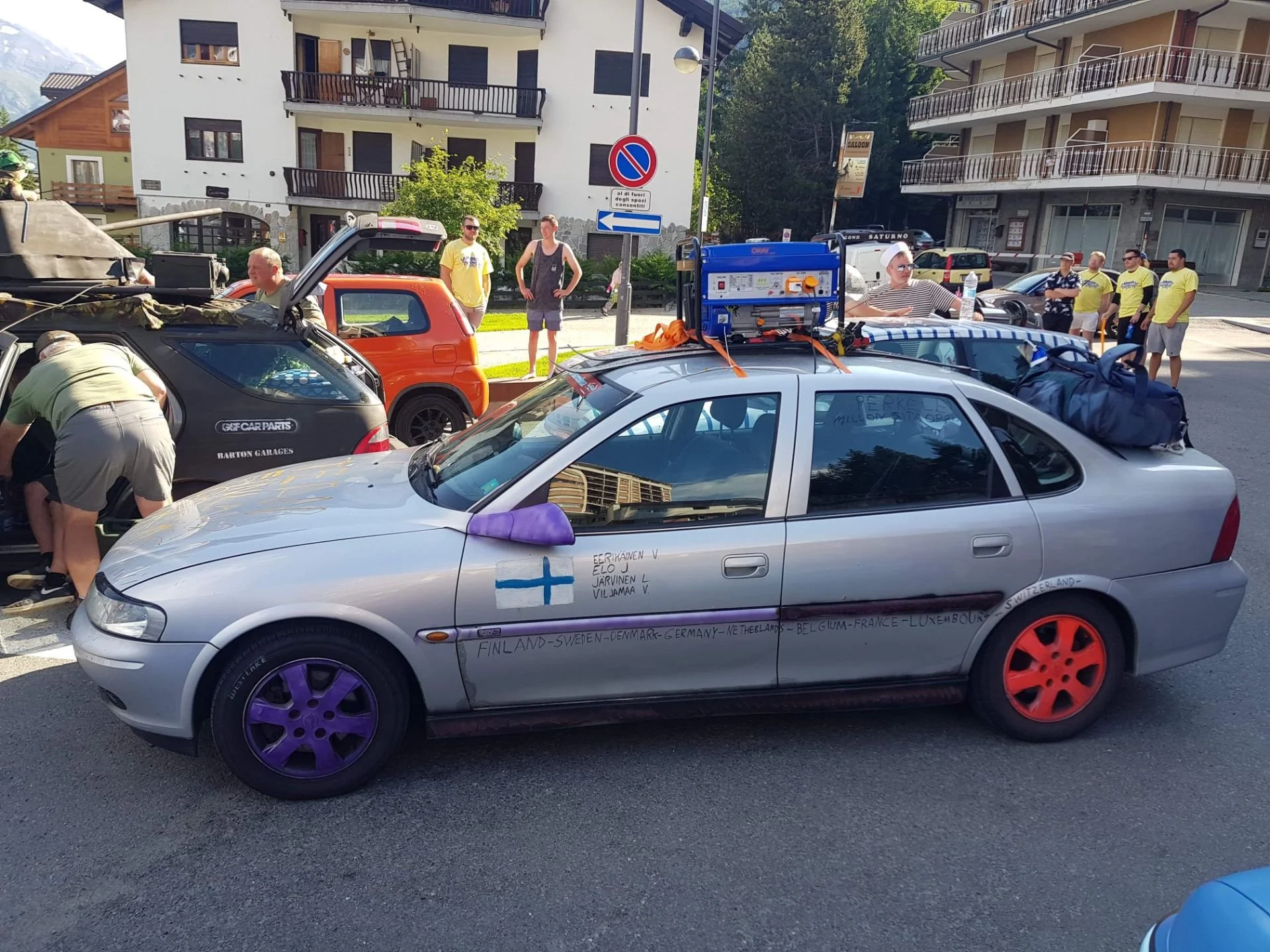A silver car with painted purple and orange wheels and handwritten text on the side, decorated with a small Finnish flag sticker, parked on a street. Several people are gathered around, some working on or near other vehicles. In the background, there