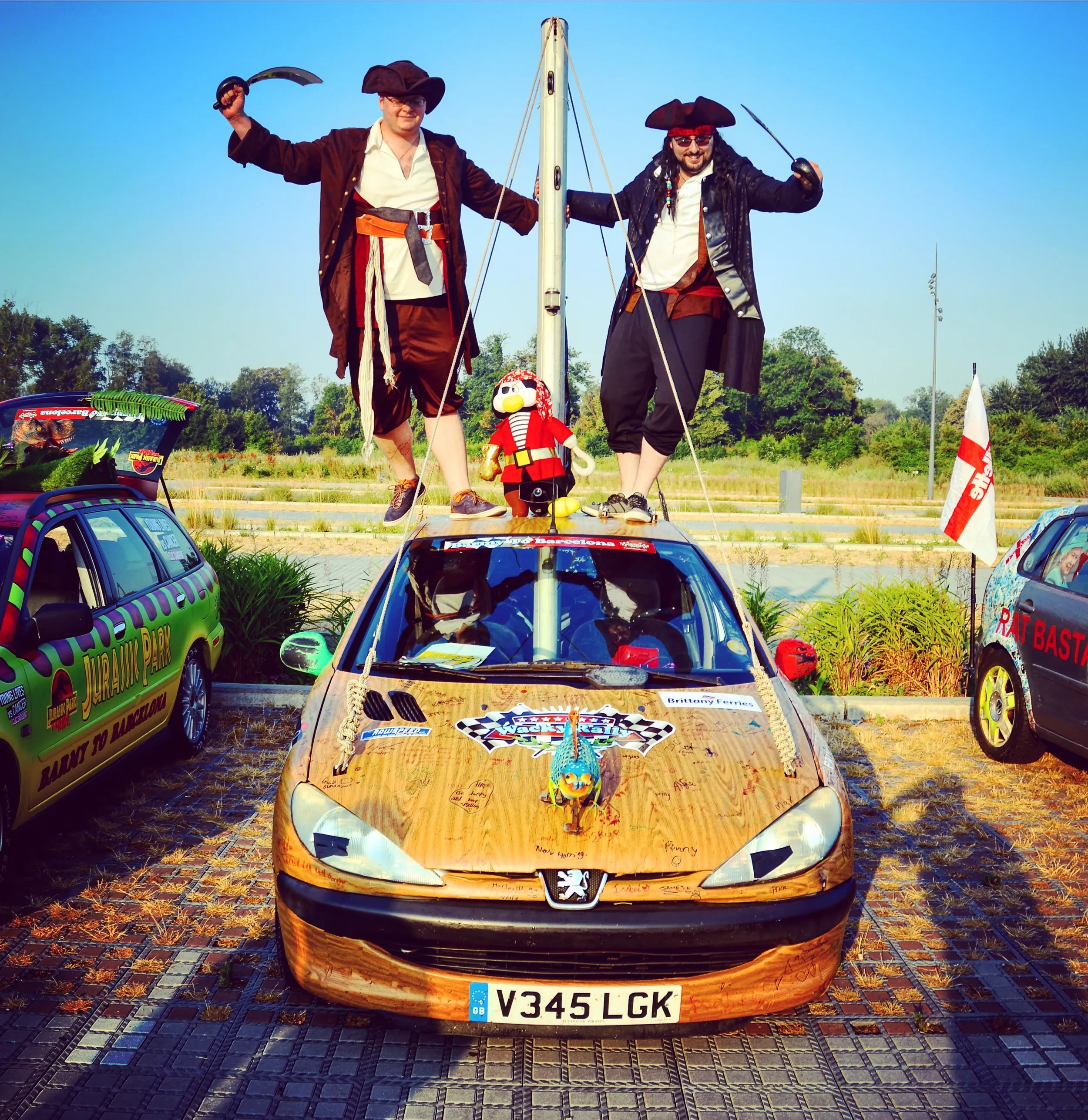 Two men in pirate costumes standing on top of a car with a pirate-themed decoration, surrounded by other decorated cars in an outdoor parking lot on a sunny day.