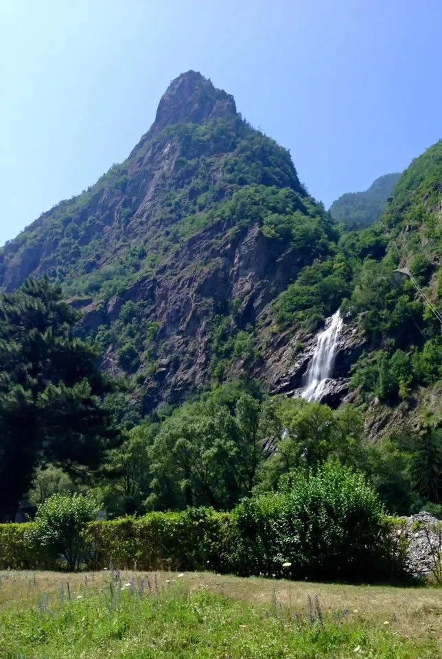 A tall, rocky mountain with a waterfall cascading down its steep side, surrounded by lush green trees and bushes under a clear blue sky.