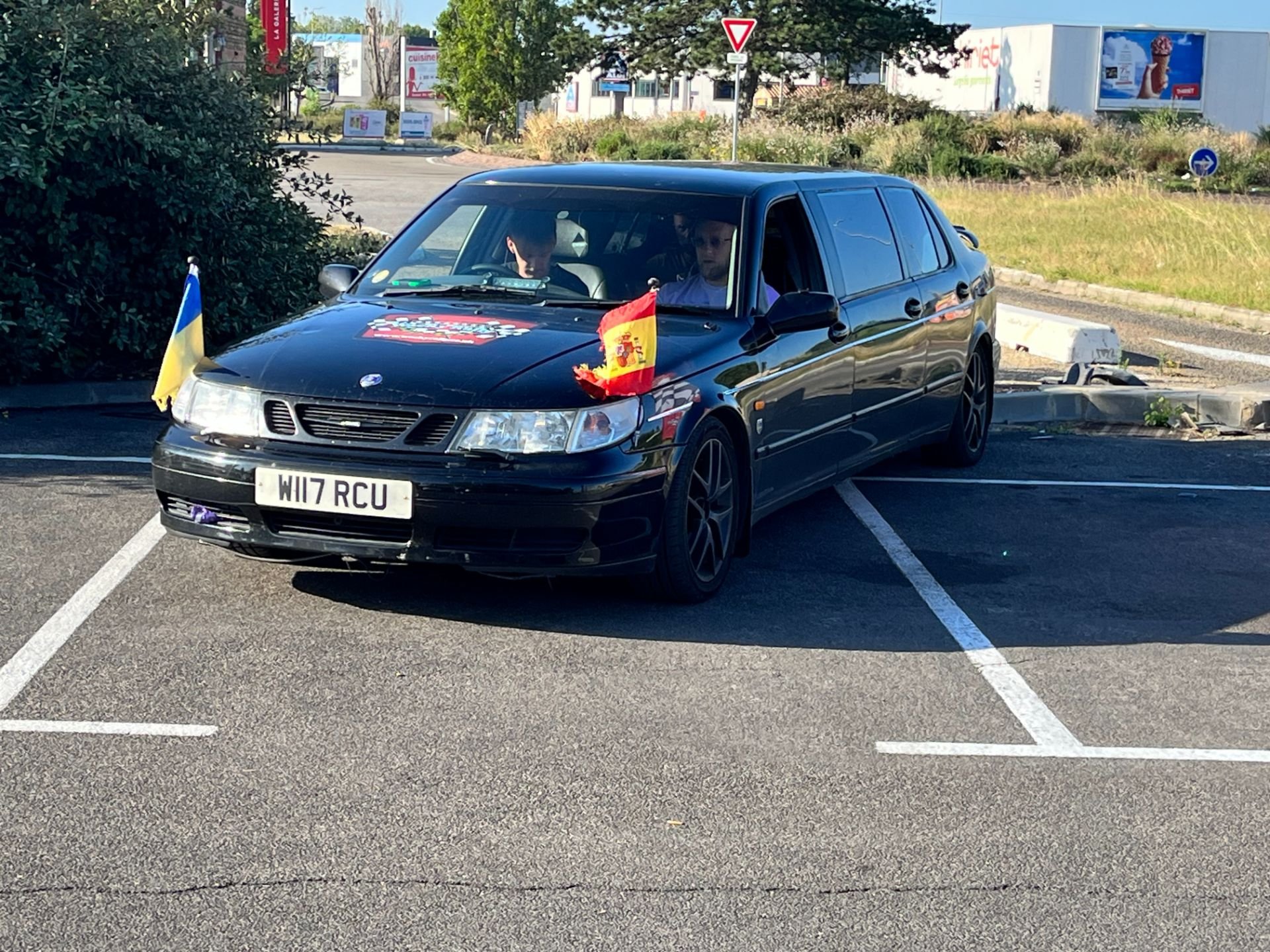 A black car decorated with flags and symbols, including the Spanish flag and the Ukrainian flag, parked in a parking lot during daytime. There are two people inside the car.